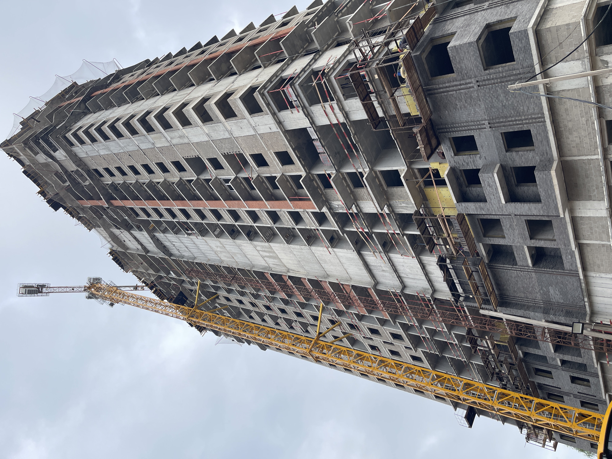 The image shows a tall building under construction in Pervomaiskii, Russia. The building's exterior is a mix of unfinished concrete and gray brickwork with rectangular window openings. A yellow construction crane stands adjacent to the building. Construction scaffolding is attached to the building. The sky is overcast. No people are visible in the frame.