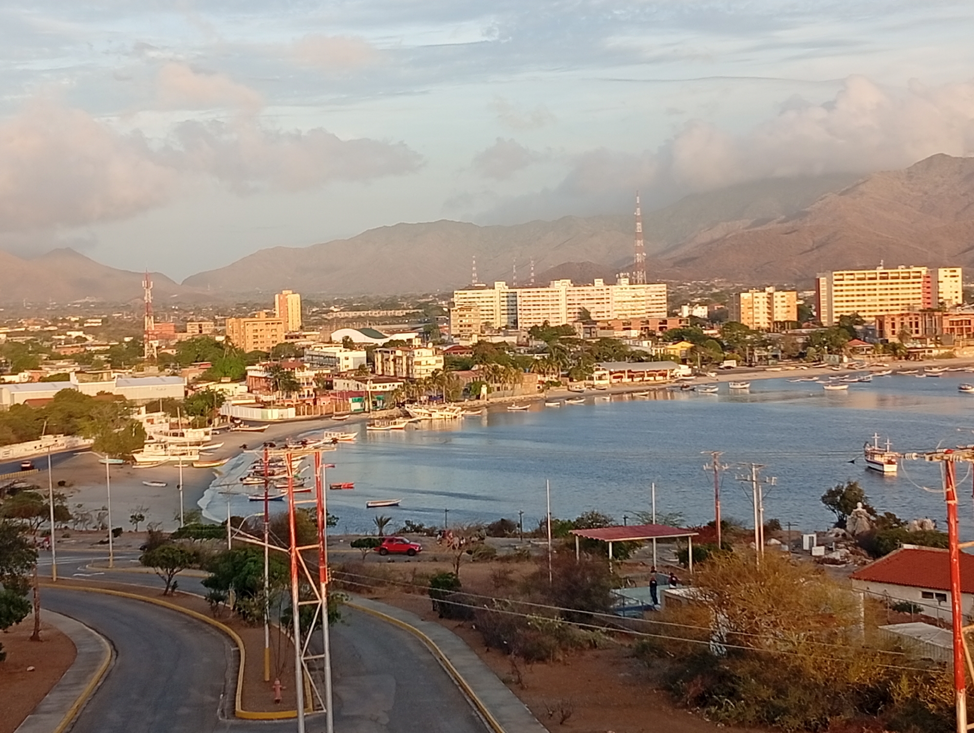 The image captures a scenic view of Juan Griego, Venezuela, overlooking a bay dotted with boats. In the foreground, a paved road with a yellow curb winds along a slight incline. Infrastructure elements, like utility poles and a red fire hydrant, are present.
Beyond the road, the bay reflects the sky, and several boats are moored or docked along the shoreline. A red car is parked near a small structure with a red roof. In the midground, numerous buildings make up the cityscape, varying in height and architectural style.
In the background, a mountain range rises against a sky with scattered clouds. Multiple transmission towers are visible amidst the urban landscape and on the mountains. The overall impression is of a coastal town nestled against a backdrop of rugged terrain.