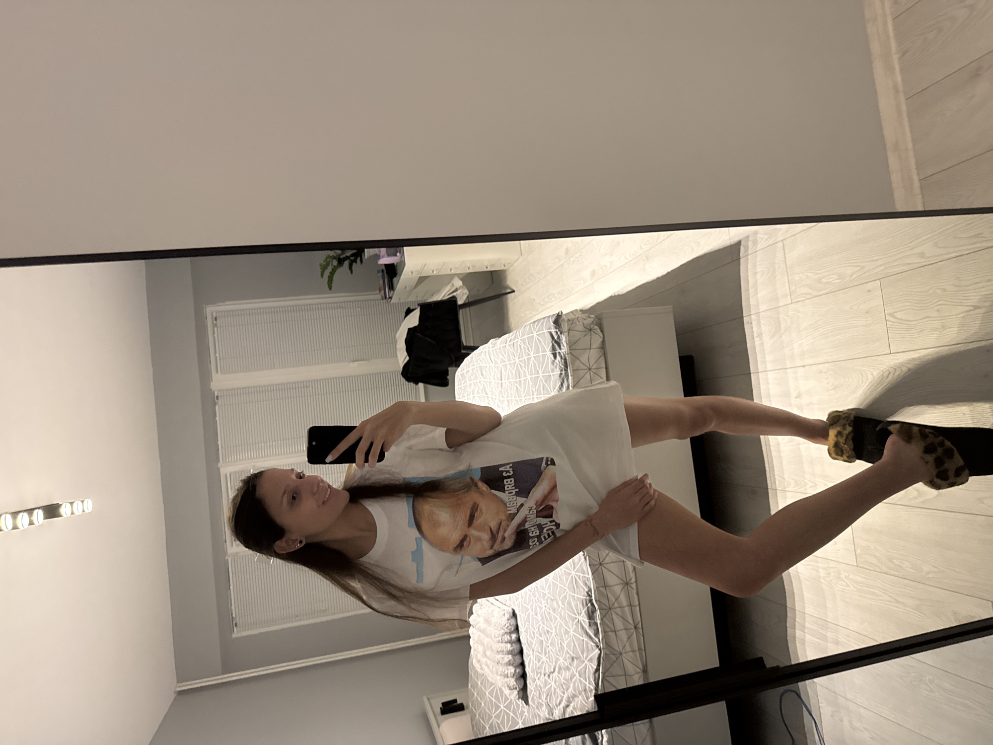 A woman is seen in a mirror selfie taken indoors. She is standing in what appears to be a bedroom, wearing a white t-shirt with a printed image on it and slippers with a leopard print pattern. The bedroom includes a bed with patterned bedding, windows with blinds, and a wall with a vanity mirror and light fixtures. The location in the city of Sofia, Bulgaria, is not identifiable from the given image.