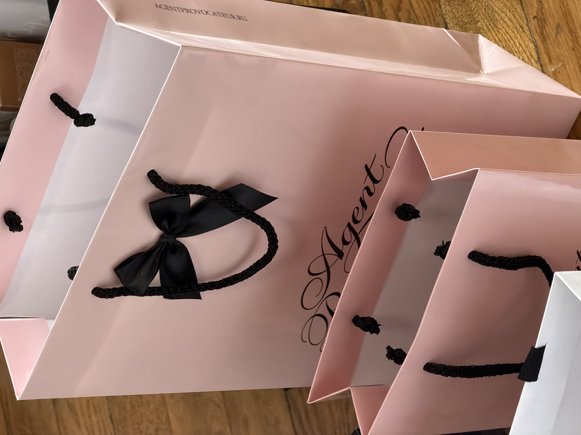 The image shows a close-up of several Agent Provocateur shopping bags on a wooden surface. The bags are pink and white with black rope handles. One bag has a black ribbon bow detail. The brand name is printed on the bags. The location cannot be determined from the image.