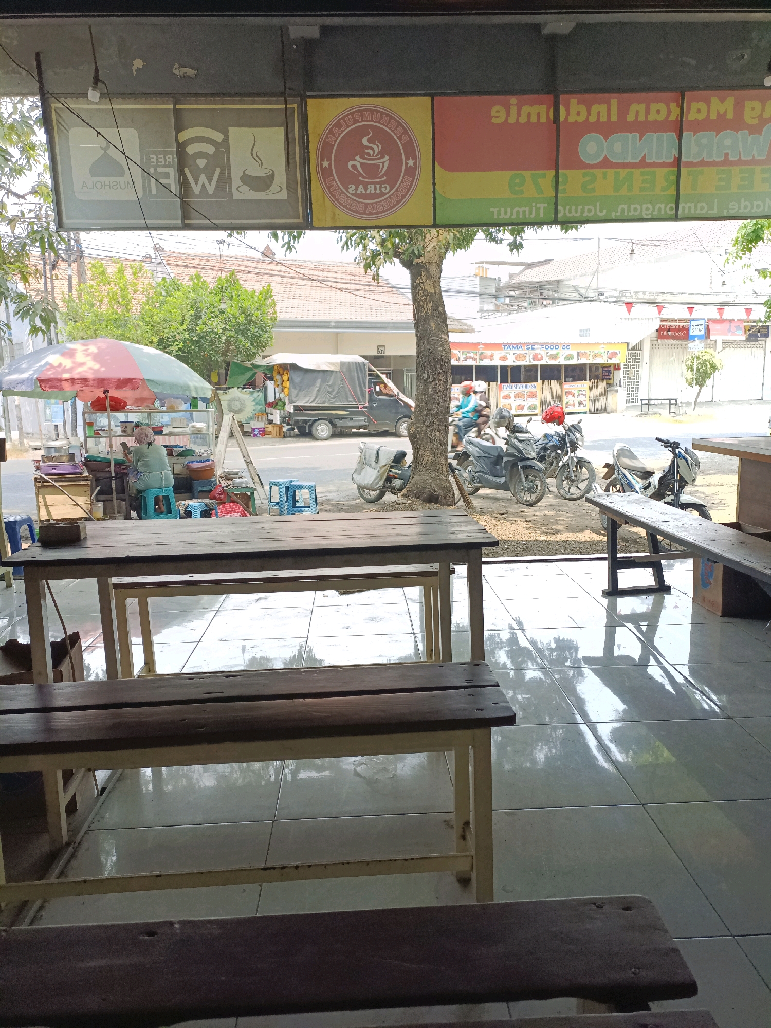 The image depicts a street-level view from the interior of a shop or restaurant in Sukomulyo, Indonesia. In the foreground, two wooden tables with metal legs are visible, one in focus and another out of focus. The floor is tiled and reflective, showing shadows from the tables and the ceiling.
Through the open doorway, a street scene unfolds. On the left, a woman sits at a food stall, tending to customers and preparing food under a colorful umbrella. The stall displays various food items and cooking equipment. Blue plastic stools are scattered around the stall.
Further down the street, several motorcycles are parked and some are in motion. One motorcycle has a red helmet on its seat. A truck with a canvas covering is parked on the right side of the road. In the background, buildings, trees, and overhead electrical wires are visible.
Above the entrance, several signs are displayed. One sign features the text "FREE WIFI" with associated icons. Another sign displays text in Indonesian and a logo with a cup of coffee. The overall impression is a bustling roadside establishment in a moderately busy Indonesian town.
