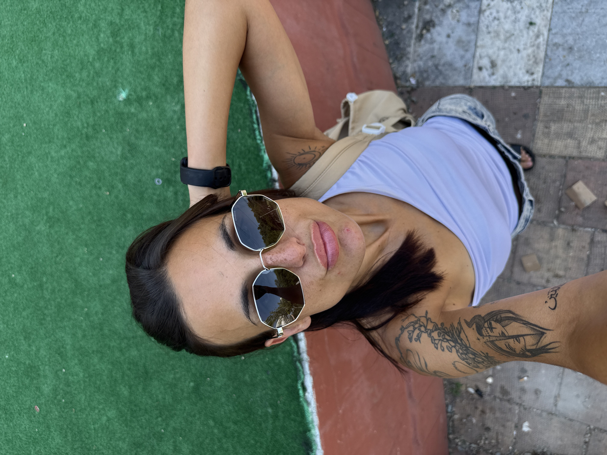 This is a high-angle, close-up, outdoor, candid shot of a woman lying on her back. She is wearing a white tank top, denim shorts, and black flip-flops. She has long, dark hair, dark eyebrows, and is wearing large, hexagonal-shaped sunglasses with a gold frame. Her lips are painted a rosy pink. She has tattoos on her arms.
The woman appears to be on artificial turf, likely a sports field or park, with a reddish-brown pathway bordering it. The ground is paved with gray and reddish-brown rectangular tiles in the background. A beige bag is slung over her shoulder.
The lighting suggests it is daytime. The woman's expression is relaxed, with her eyes closed or looking down. There is no visible text in the image. The setting appears to be urban.