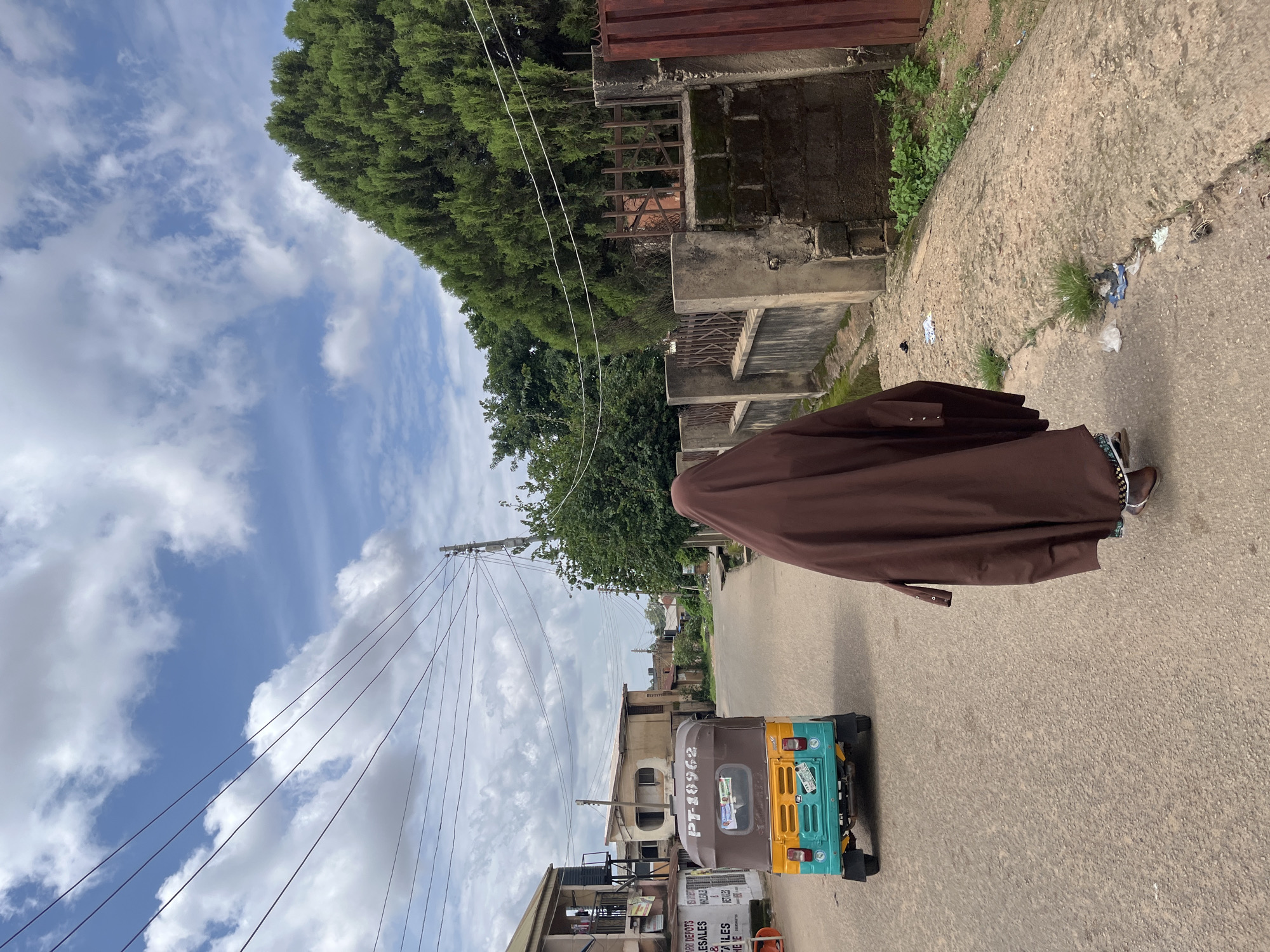A person wearing a full brown abaya walks away from the camera down a paved road in Jos, Nigeria. They are walking in the middle of the road, away from a parked, colorful auto-rickshaw. The auto-rickshaw is teal, yellow, and brown, with "PT-18962" printed on the back. The person is partially obscuring a large green tree and a concrete fence on the right side of the road. The sky is blue with scattered white clouds. Power lines are visible running across the sky. Buildings are on the left side of the road.