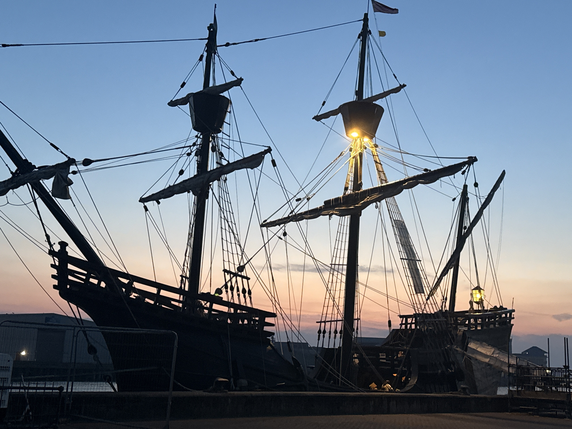 The image depicts a historical sailing ship, likely a replica, docked at dusk in Great Yarmouth, United Kingdom. The ship features multiple masts with rigging and sails furled. A bright yellow lantern is illuminated on the deck of the ship, casting a warm glow. The sky displays a gradient of blues and soft pinks, indicating the transition from day to night. In the background, silhouetted industrial buildings and structures are visible. The ship is positioned on a quayside, with a safety fence in the foreground. A few figures are dimly visible on the deck of the ship, engaged in what appears to be maintenance or preparation. The overall scene evokes a sense of maritime history and the enduring appeal of traditional sailing vessels.