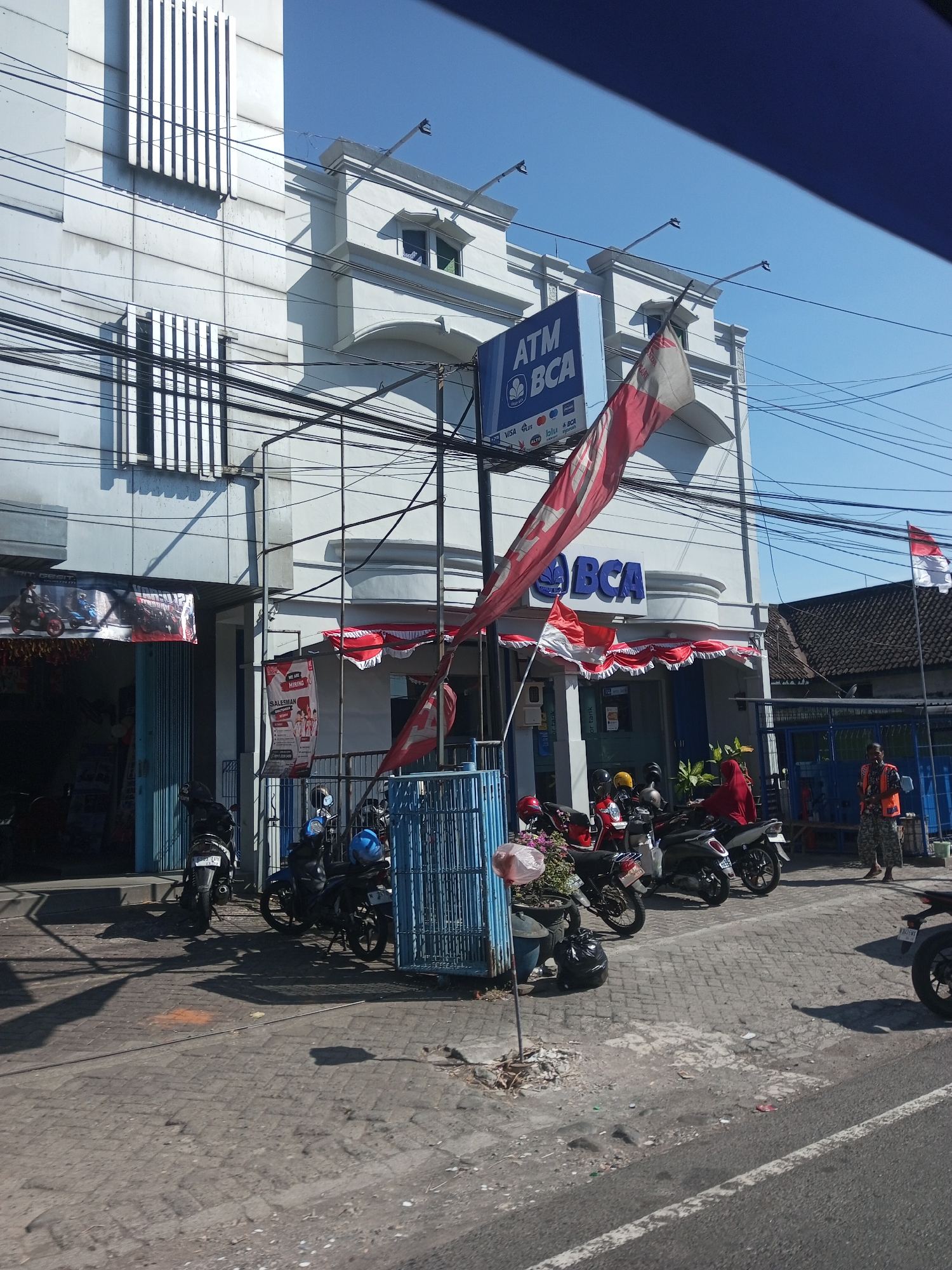 The image depicts the exterior of a Bank Central Asia (BCA) branch and ATM located on a street in Purwosari, Indonesia. The building is white with architectural detailing, including decorative window frames. Several motorcycles are parked along the sidewalk in front of the bank. A blue metal gate is visible on the left side of the bank entrance. Red and white banners and flags, consistent with Indonesian national colors, are displayed along the front of the building. A prominent ATM sign is mounted above the entrance, and a red and white flag is draped over a pole in front of it. A person wearing an orange vest and patterned pants stands to the right, near a blue structure. Another individual in a red head covering is visible near the motorcycles. Electrical wires are strung across the scene. The ground is paved with bricks, and a paved road is in the foreground. The sky is clear and blue.