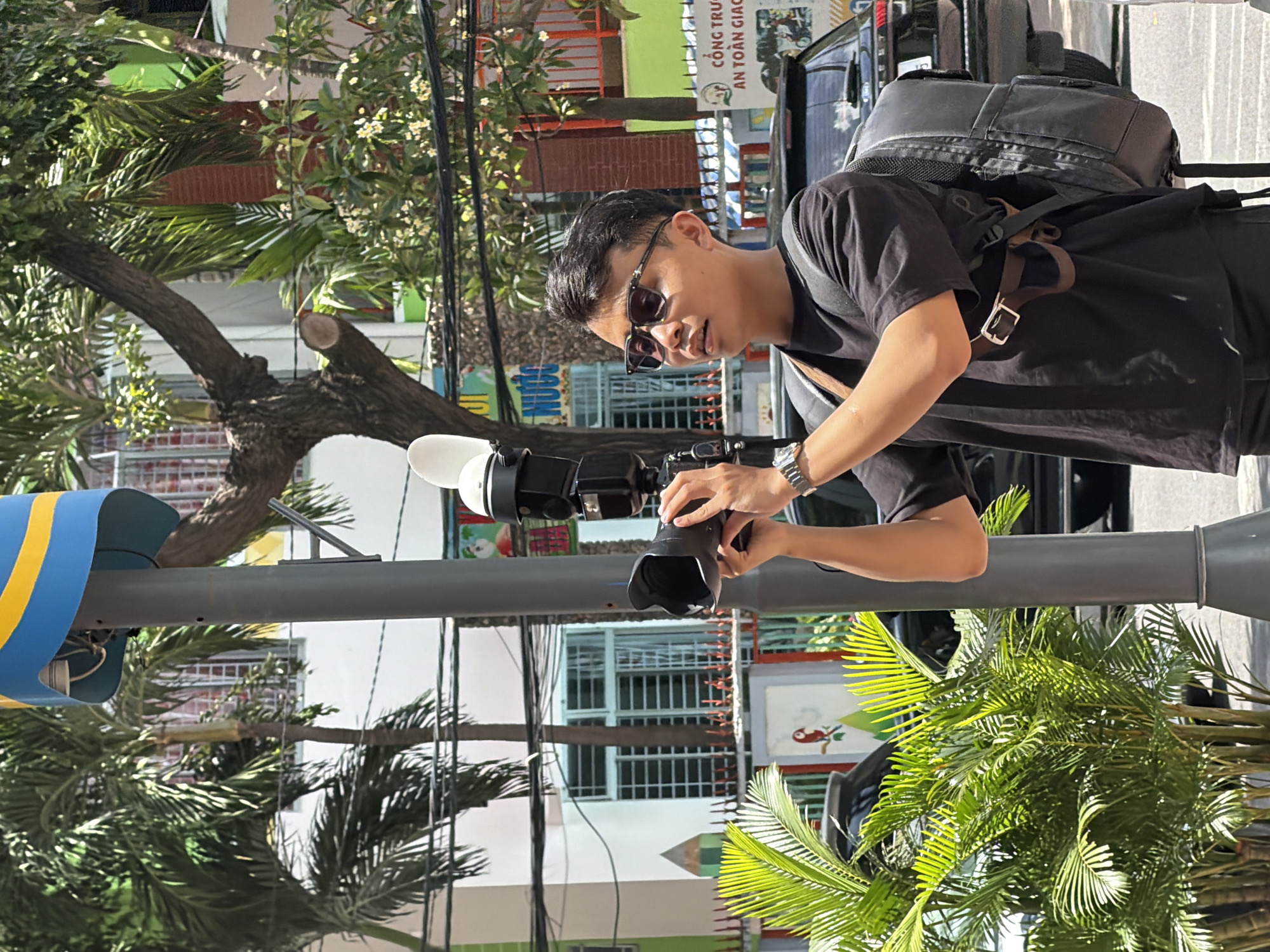A young man, wearing a black t-shirt, sunglasses, and a backpack, is standing next to a lamppost and holding a professional camera with a flash attachment. He appears to be taking a picture. The background suggests an urban environment in Nha Trang, Vietnam, with palm trees, buildings with colorful facades, and parked vehicles. One of the buildings has signage indicating "Công Trunc An Toàn Giao" which translates to "Traffic Safety Center". The overall scene depicts a street on a sunny day.