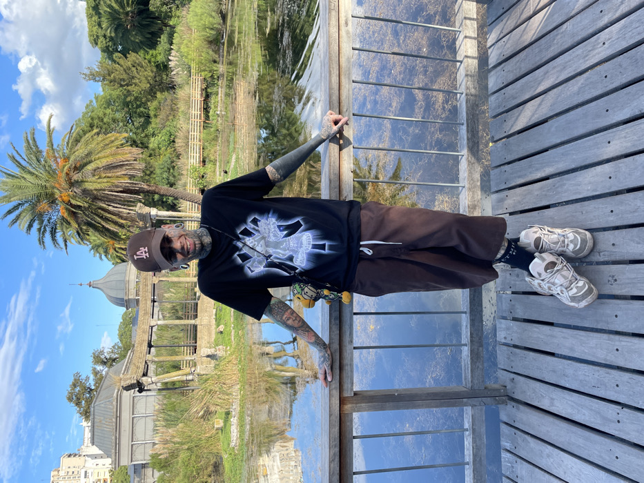 A tattooed person stands on a wooden boardwalk next to a clear water canal, reflecting the surrounding park. The person wears a black graphic t-shirt, brown shorts, sneakers, a Los Angeles baseball cap, and carries a small, character-themed shoulder bag. Trees, including a palm, and a glass-domed structure are visible in the background. The sky is partly cloudy. The image is rotated to the left.
