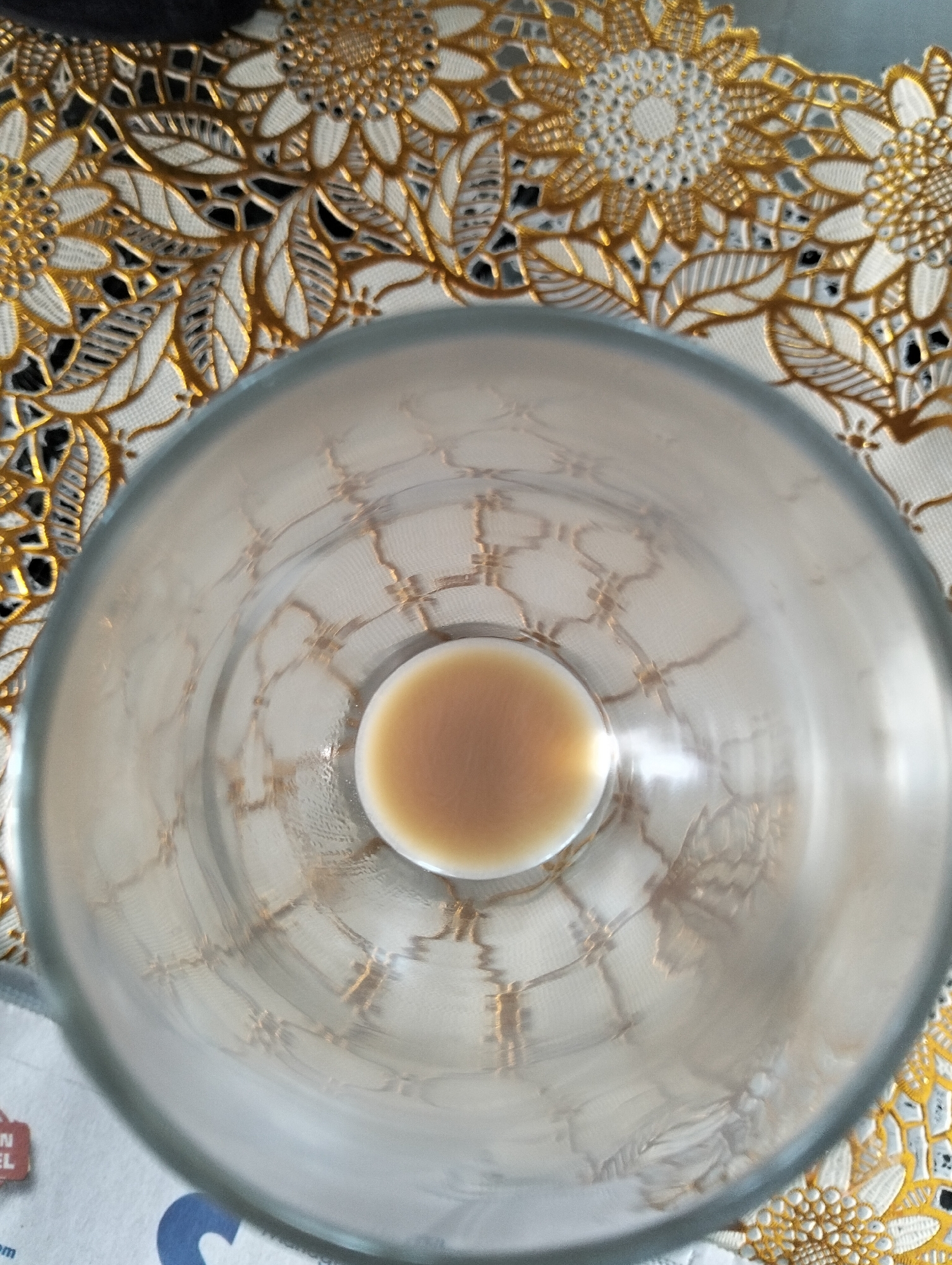 This is a close-up, top-down shot of a clear glass with a small amount of light brown liquid at the bottom. The glass is resting on a table covered with a white tablecloth that features an ornate gold pattern of sunflowers and leaves. The pattern is intricate and appears to be embroidered or appliquéd.
The lighting suggests an indoor setting, possibly in the daytime due to the ambient light reflecting off the glass. There are no people visible in the image. The focus is on the glass and the tablecloth. The glass itself has some subtle circular etchings or a pattern visible on its interior surface, reflecting the light. The liquid within the glass is a uniform light brown, suggesting it could be coffee, tea, or a similar beverage.
There is some visible text in the lower left corner, partially obscured by the glass and tablecloth. It appears to be from some printed material, possibly a newspaper or packaging, with the letters "EL" and "om" being the most legible.
The overall impression is one of a simple, domestic scene, perhaps a moment of pause with a drink. The decorative tablecloth adds a touch of elegance to the otherwise plain setting. The location context of Julicotmeurak, Indonesia, does not provide specific visual cues within the image itself.