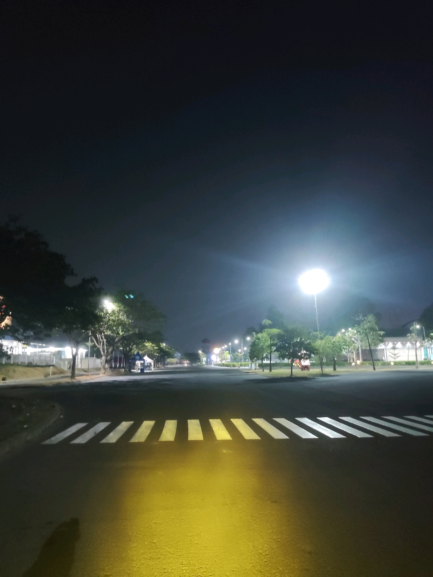 The image depicts a nighttime scene at a pedestrian crossing on a road in Telagamurni, Indonesia. A bright street light illuminates the area, casting a yellow glow on the asphalt and the white stripes of the crosswalk. Trees line the roadside, and some buildings and tents are visible in the background. The sky is dark, with no stars or moon visible. The overall impression is one of a quiet, well-lit urban environment at night.