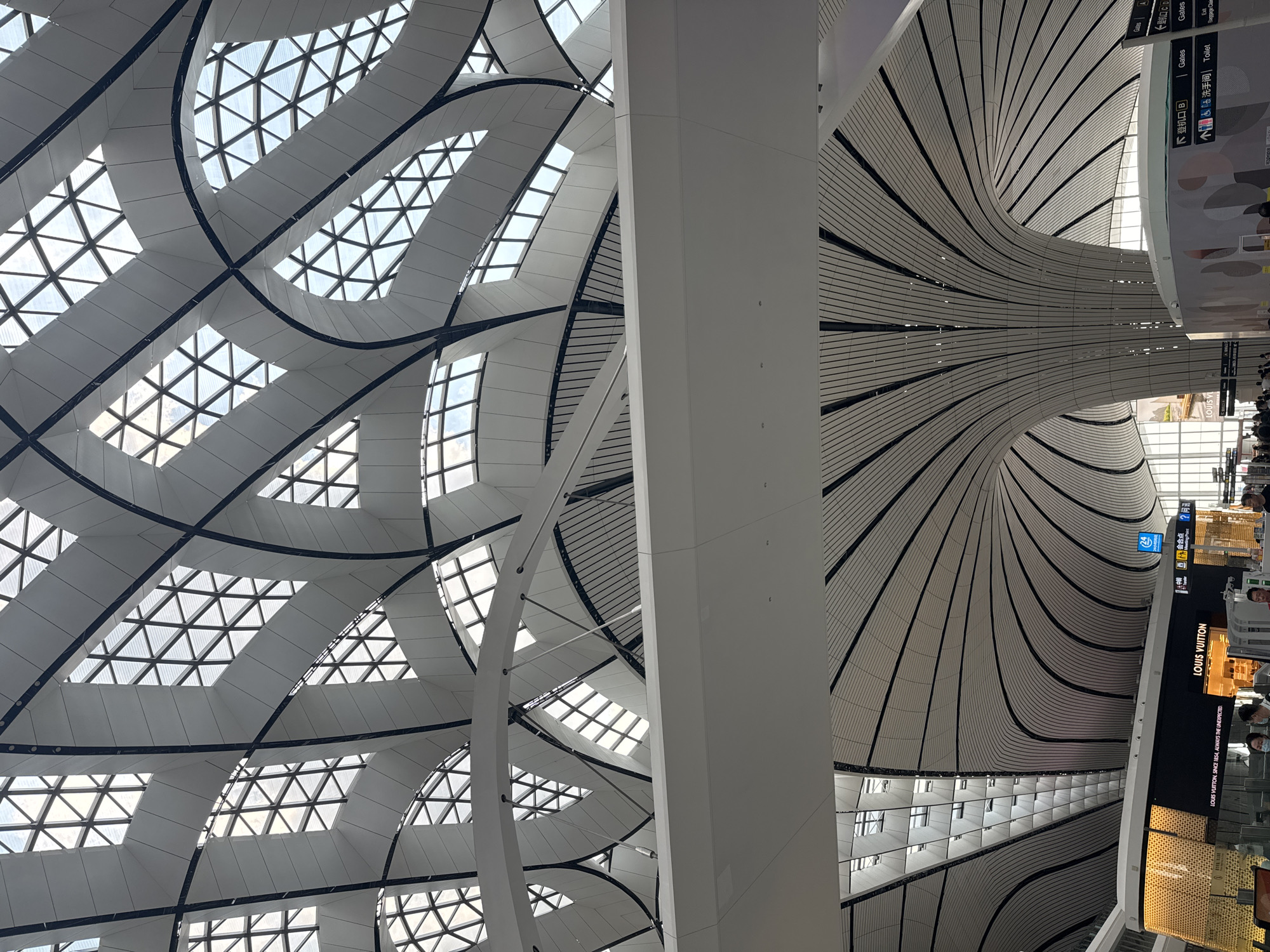 The image shows the interior of Beijing Daxing International Airport, located in Guangyang District, China. The airport features a distinctive, large, and modern architectural design, with a prominent undulating ceiling composed of white, curved panels and dark structural beams. The ceiling is punctuated by triangular skylights that allow natural light to enter the space. In the lower portion of the image, a Louis Vuitton store is visible, with its signage and illuminated storefront. People are seen walking around the airport concourse, some wearing masks. The overall environment is clean, spacious, and brightly lit due to the natural light from the ceiling.