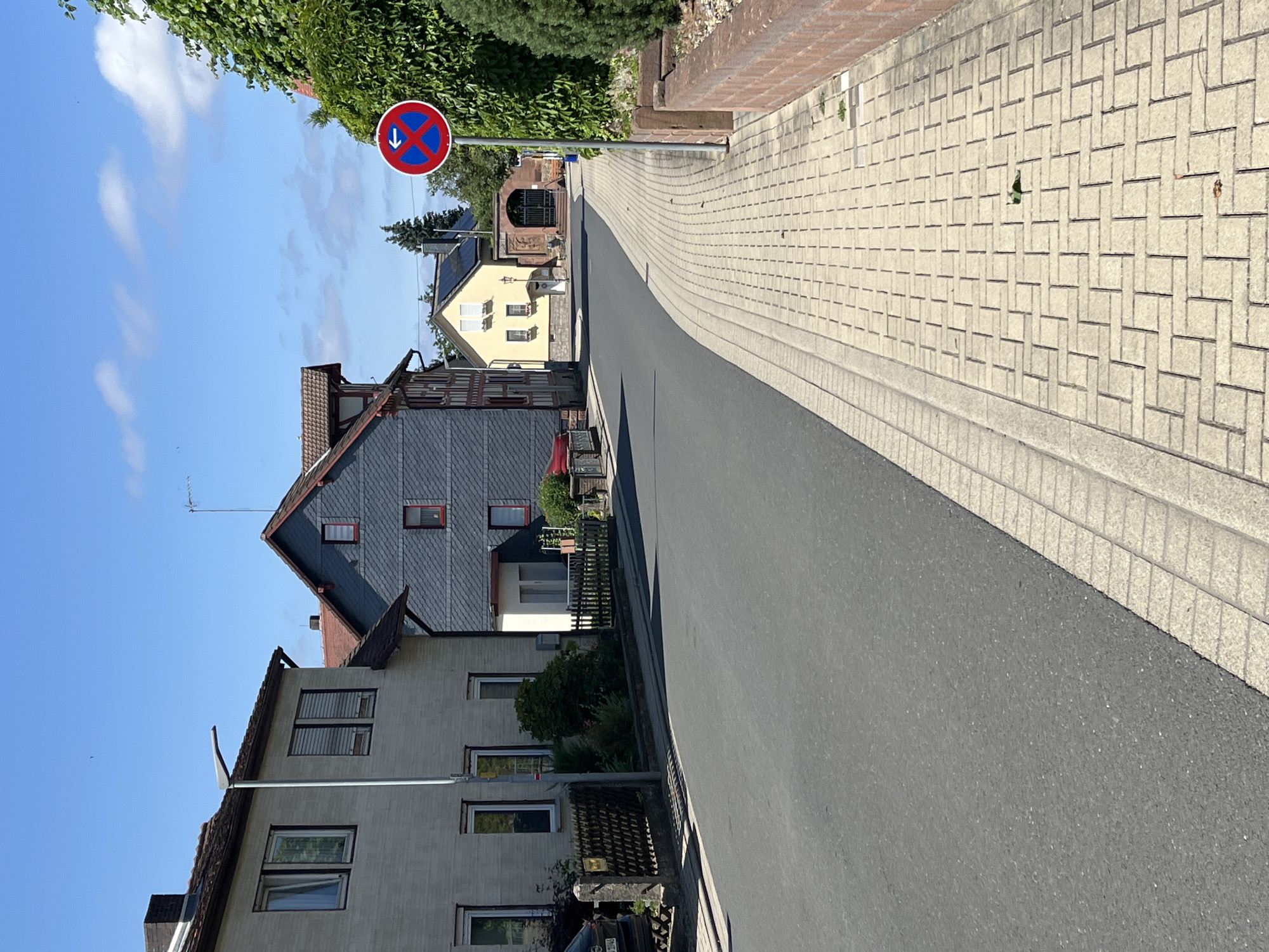 The image depicts a street scene in Neustadt, Germany, under a clear blue sky with scattered white clouds. On the left side of the street, a light-colored, multi-story building with several windows is visible. A lamppost stands in front of this building. Further down the street, a darker, slate-clad house with red window frames is situated. A bright yellow house with white-framed windows is visible in the distance, adjacent to a building with a large arched entryway.
On the right side of the street, a paved sidewalk made of light-colored rectangular bricks runs alongside the road. A red and blue traffic sign with a white arrow pointing left and a diagonal red line across a blue circle indicates a no-parking zone for vehicles to the left. Adjacent to the sidewalk, a low brick wall and lush green foliage are present. The road surface appears to be asphalt. There are no people or vehicles actively engaged in any actions within the frame.