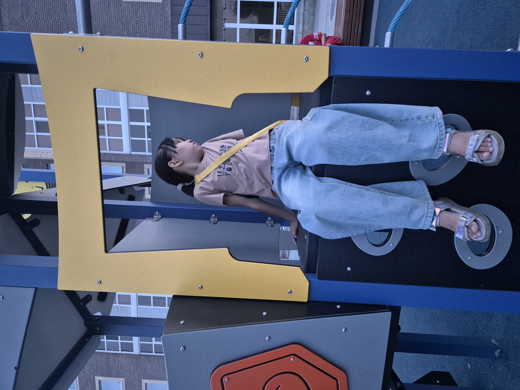 A young girl sits on a playground structure, looking to her right. She is wearing a light brown t-shirt with text, light blue baggy jeans, and sparkly sandals. A yellow strap is visible across her chest. The playground structure is made of brightly colored panels, including yellow, blue, and gray, with some red accents. In the background, apartment buildings with visible windows are present, suggesting an urban environment. The scene appears to be an outdoor playground. The image was taken in Kokshetau, Kazakhstan.