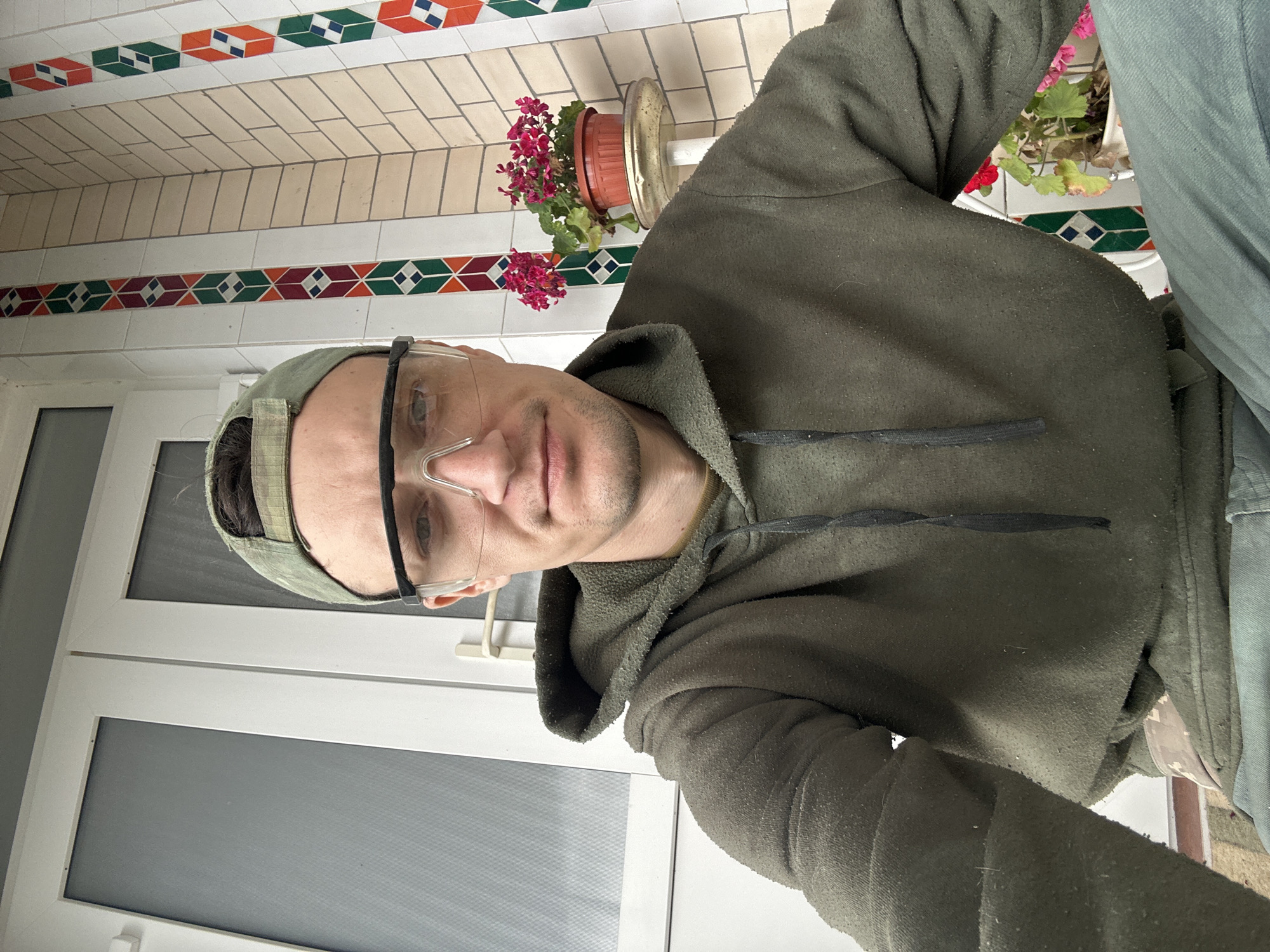A man wearing a green hoodie, a camouflage baseball cap, and clear safety glasses is taking a selfie. He is outdoors, possibly on a porch or patio, with a white door and decorative tilework in the background. A potted plant with pink flowers is visible to his right. The man appears to be looking directly at the camera. The scene suggests a casual or potentially work-related setting.