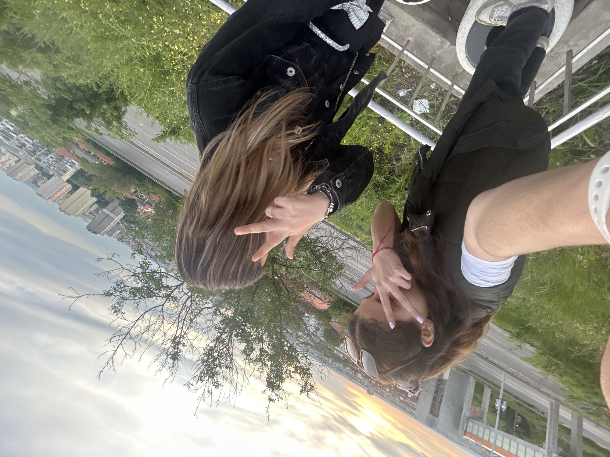 Two young women are positioned on a walkway, likely a bridge or elevated platform, overlooking a cityscape. One woman has long brown hair and wears a black denim jacket, standing to the left of the frame. The other has dark hair, wears sunglasses, and is dressed in a black vest over a white shirt. Both are making hand gestures with their fingers, pointing downwards.
The cityscape in the background is suggestive of Ruse, Bulgaria, due to its architecture, which includes apartment buildings and other urban structures. The sky is transitioning from day to night, evidenced by a distinct sunset with orange and yellow hues.
The walkway has metal railings and concrete or stone beneath the women's feet. Patches of grass are visible surrounding the structure, suggesting an outdoor environment. One person's arm extends into the frame, indicating the photo is a selfie.