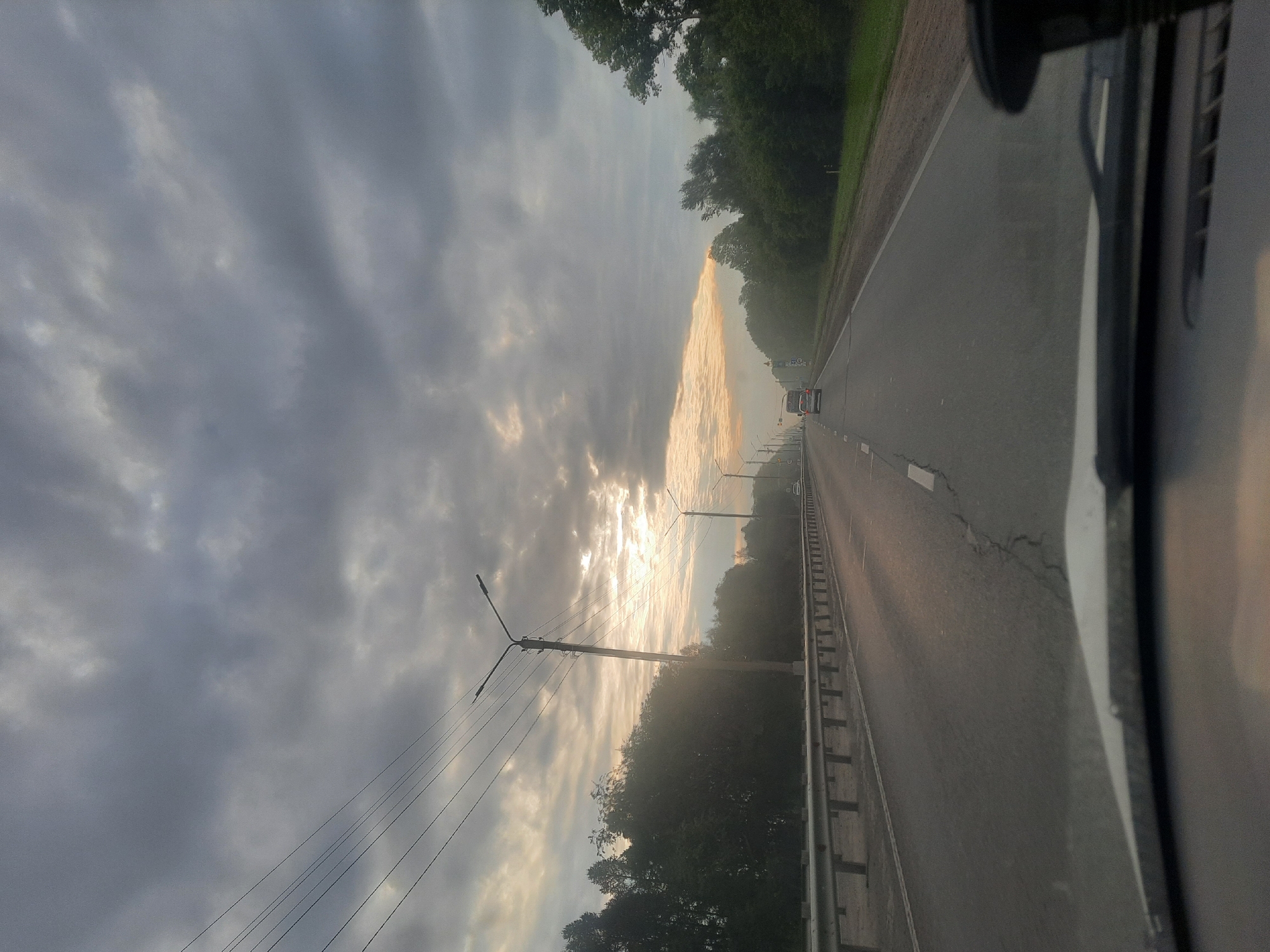 The image depicts a view from inside a vehicle looking out at a highway in Barnaul, Russia. The sky is heavily overcast with dramatic grey clouds, but a sliver of sunlight breaks through near the horizon, creating a bright, hazy band. The highway stretches into the distance, with a metal guardrail lining the left side. Trees and vegetation line the sides of the road, appearing dense and green. Several streetlights are visible along the highway, and a few vehicles are present, including a white car in the distance and a darker vehicle further ahead. The road surface shows some cracking and wear. The foreground of the image is dominated by the dashboard and windshield of the vehicle.