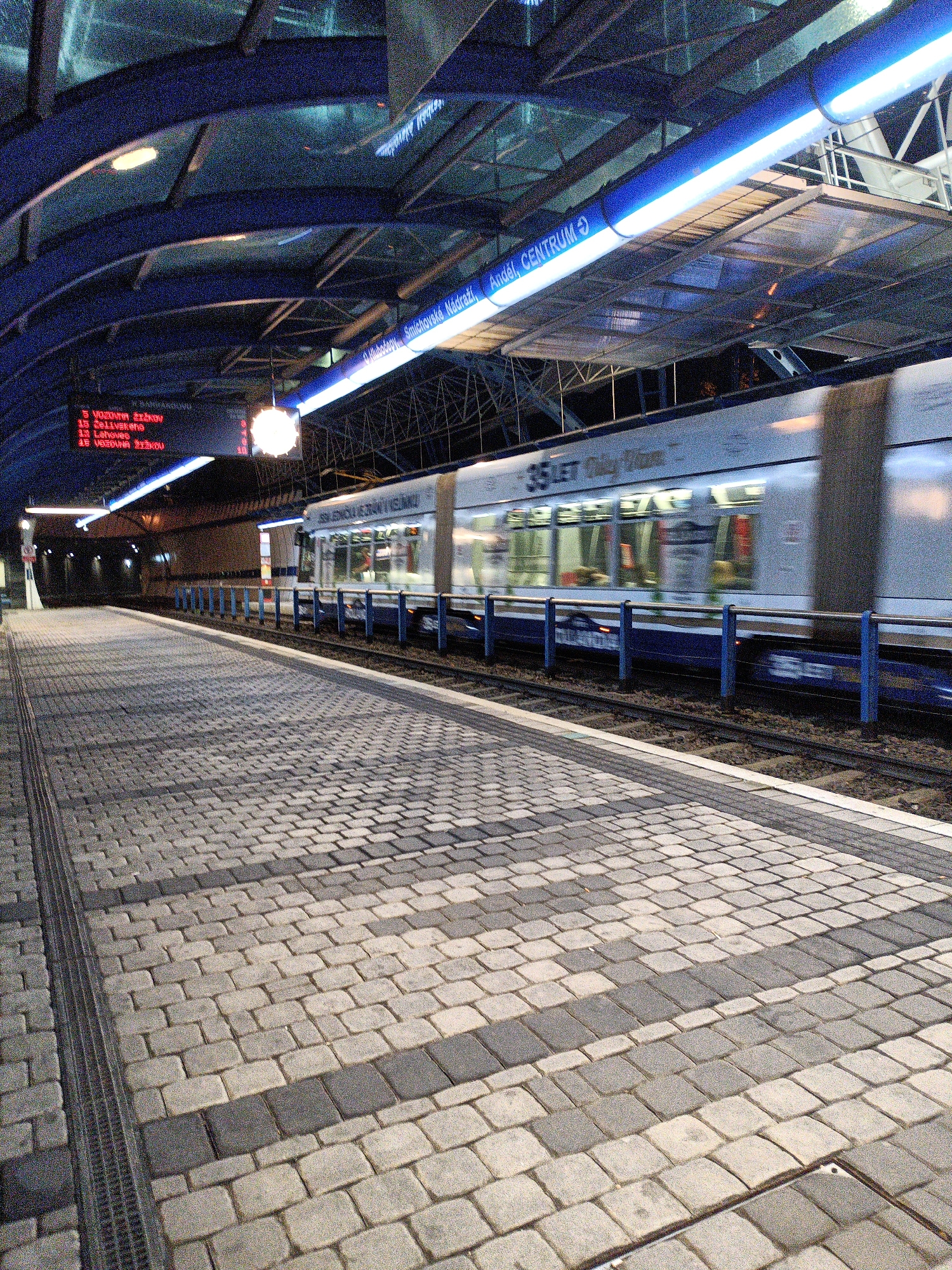 The image depicts a modern, covered tram station platform in Prague, Czechia. The platform is paved with gray and darker gray interlocking bricks, with a metal grate running along the left edge. A tram is moving rapidly on the tracks to the right, its motion creating a blur. The tram is white and blue, with visible windows. Above the platform, a curved, dark blue roof structure is supported by beams. Blue illuminated signage runs along the edge of the roof, indicating destinations. One sign clearly reads "Smíchov, nádraží" and "Anděl, Centrum". A digital display board with red text is also visible, showing tram numbers and destinations. The lighting is artificial, with bright blue lights integrated into the roof structure and a circular lamp hanging from the ceiling. The overall impression is of a functional and contemporary urban transportation hub.