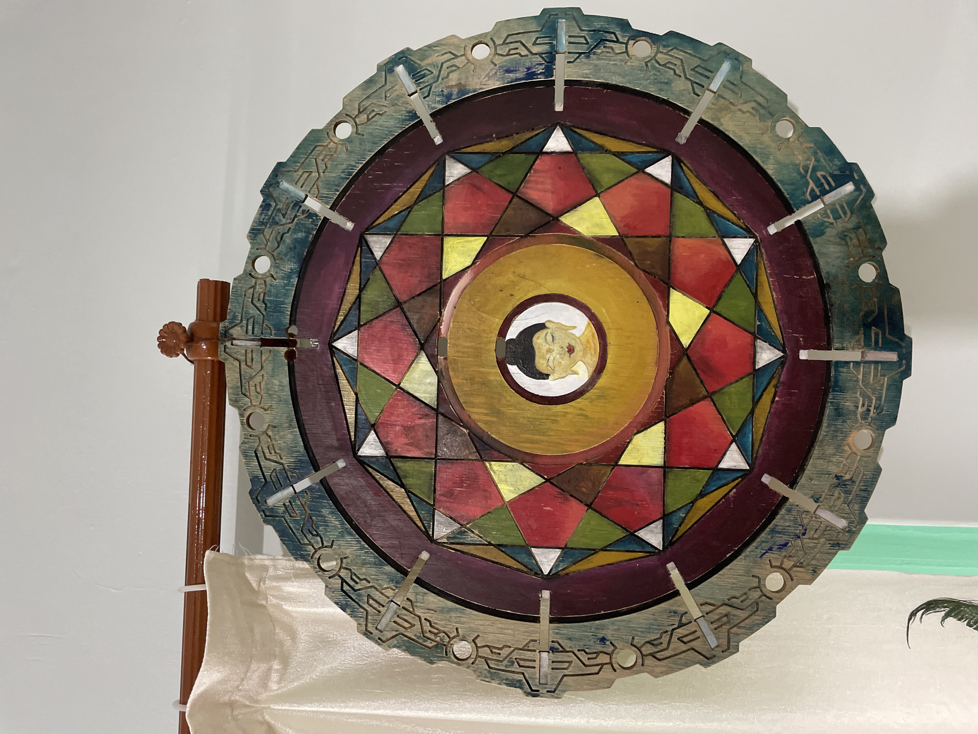 A circular, mandala-like artwork with a depiction of Buddha's face at its center hangs on a white wall in Bhagsu, India. The artwork is constructed from wood and features intricate geometric patterns in vibrant colors like red, yellow, green, and purple. The outer rim of the mandala is adorned with carved designs and metallic elements. The artwork is suspended by a wooden rod with decorative carvings, which is supported by a white bracket. A light-colored curtain is visible to the left of the artwork, partially obscuring the wall. The lighting in the scene is soft, suggesting an indoor setting.