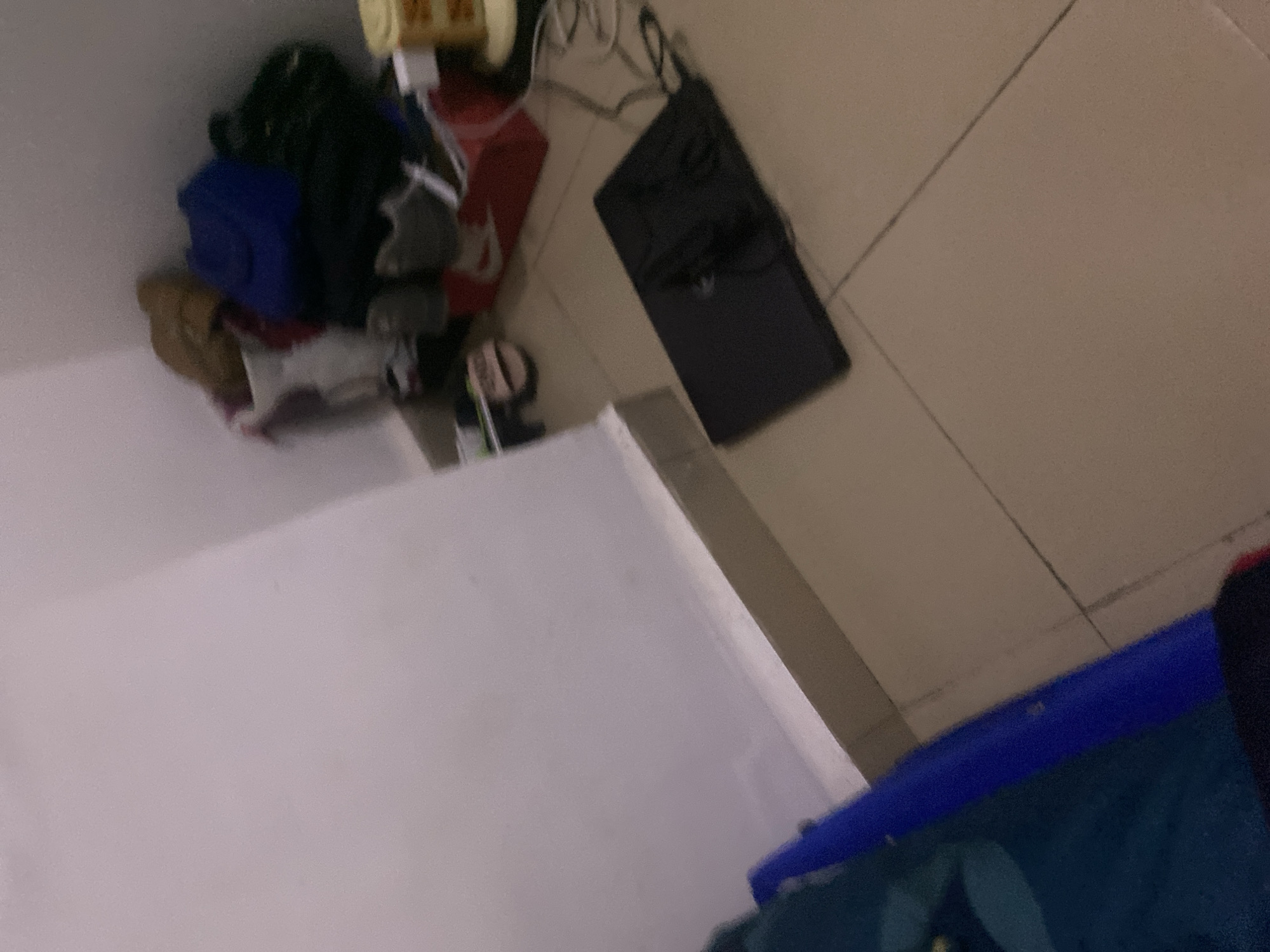 The image depicts a cluttered corner of a room. A pile of items, including bags and possibly shoes, is stacked in the far right corner. In the foreground, a black laptop is placed on the tiled floor, with cables extending from it. To the left, a blue object, possibly a bed or a piece of furniture, is partially visible. The walls are white, and the floor is tiled in a beige or light brown color. The scene appears to be an indoor residential space. There are no people visible in the image. The overall impression is one of disarray and a lived-in environment.