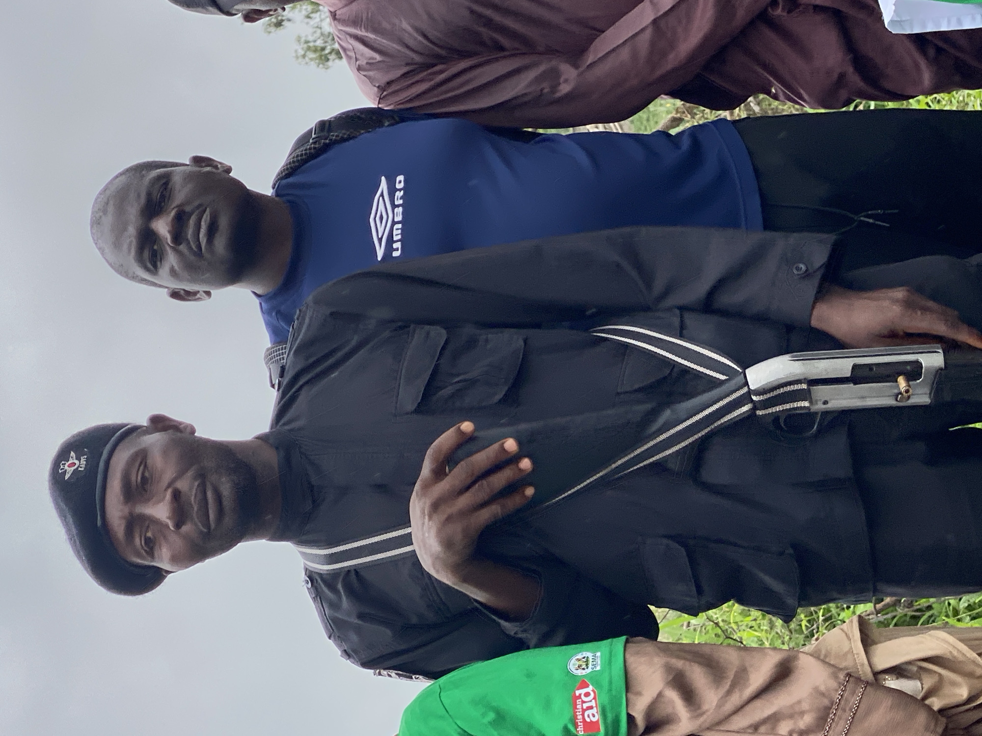 In the city of Kawo, Nigeria, four individuals stand outdoors. The person in the foreground, on the left, is a Black male wearing a black beret with a badge, a black long-sleeved uniform with pockets, and black pants. A silver shotgun is slung across his chest with a white and grey strap. His right hand rests on his left forearm. To his right stands another Black male wearing a blue t-shirt with the Umbro logo and black pants. Behind them, a third Black male is partially visible, wearing a dark colored t-shirt. To the far left of the frame, another individual is visible wearing a green t-shirt with the "Christian Aid" logo and a tan garment. The background appears to be an outdoor environment with greenery and a cloudy sky.