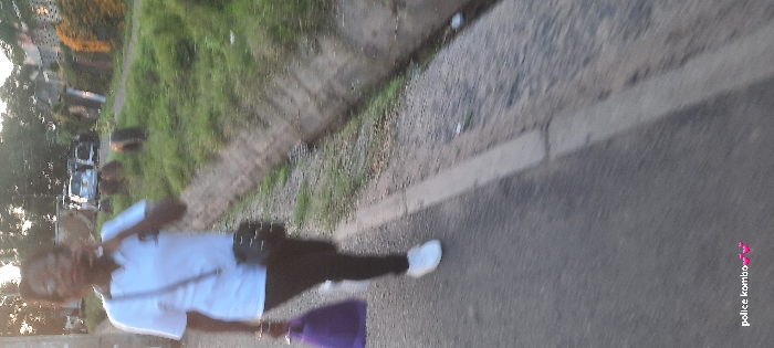 The image captures a person walking along a paved road in what appears to be an urban or semi-urban environment during the late afternoon or early evening, possibly in Nairobi, Kenya. The overall image quality is blurry, suggesting either motion blur or a low-resolution capture.
The central subject is an individual, seen from a slightly elevated perspective and behind, walking on the right side of the road. They are dressed in a white short-sleeved top, dark trousers or leggings, and white sneakers. A dark shoulder or crossbody bag is visible across their body. In their left hand, they hold a distinct purple object, possibly a small bag or an umbrella. The person's face and upper body are largely indistinct due to the blur.
The setting includes a paved asphalt road on the right, bordered by a concrete curb. To the left of the curb, there is a grassy verge with green vegetation and what appears to be a shallow ditch or embankment running parallel to the road. In the background, further down the street, several vehicles are faintly visible, including at least one white car. Distant buildings and trees can be discerned on the far left horizon. The lighting indicates the golden hour, providing a soft, warm glow across the scene. A small watermark with the text "police kombo" accompanied by pink heart symbols is present in the lower right corner of the image.