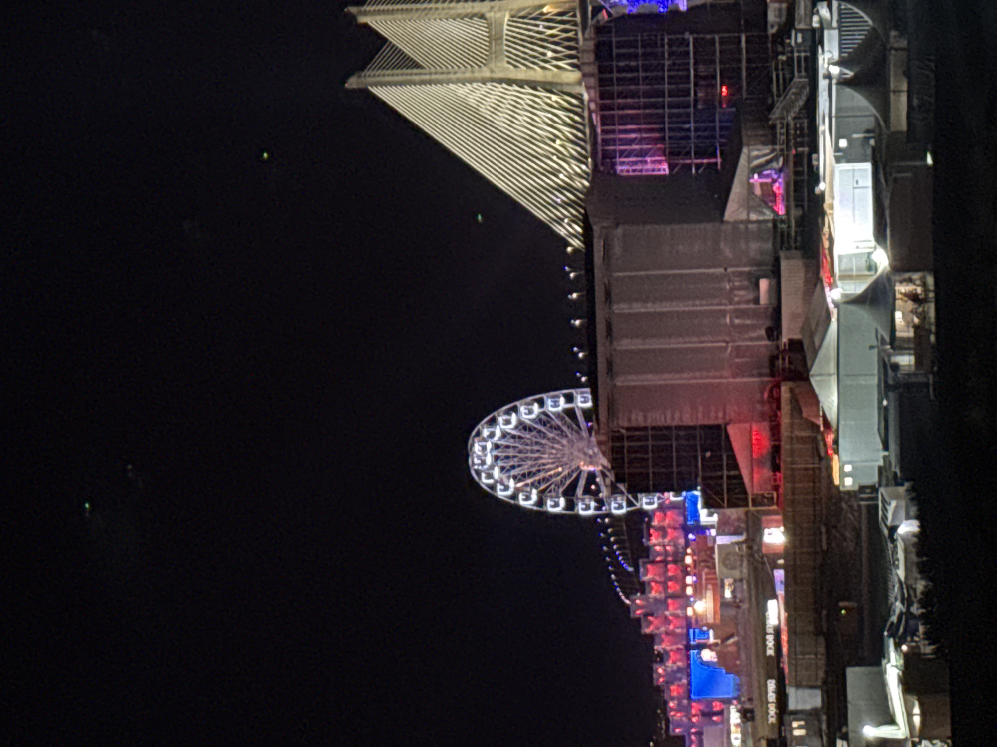 The image depicts an outdoor event at night, likely a festival or concert, held near the Vasco da Gama Bridge in Loures, Portugal. A large Ferris wheel, illuminated with white lights, is prominently visible on the left side of the frame. The Vasco da Gama Bridge itself, a cable-stayed bridge, is illuminated with white lights, highlighting its distinctive architecture. In the foreground, various temporary structures, including tents and stages, are set up. Some of these structures are also lit, casting a warm glow on the scene. The overall atmosphere appears festive and lively, with the bright lights contrasting against the dark night sky. There are a few faint green lights visible in the sky, possibly drones or distant aircraft.