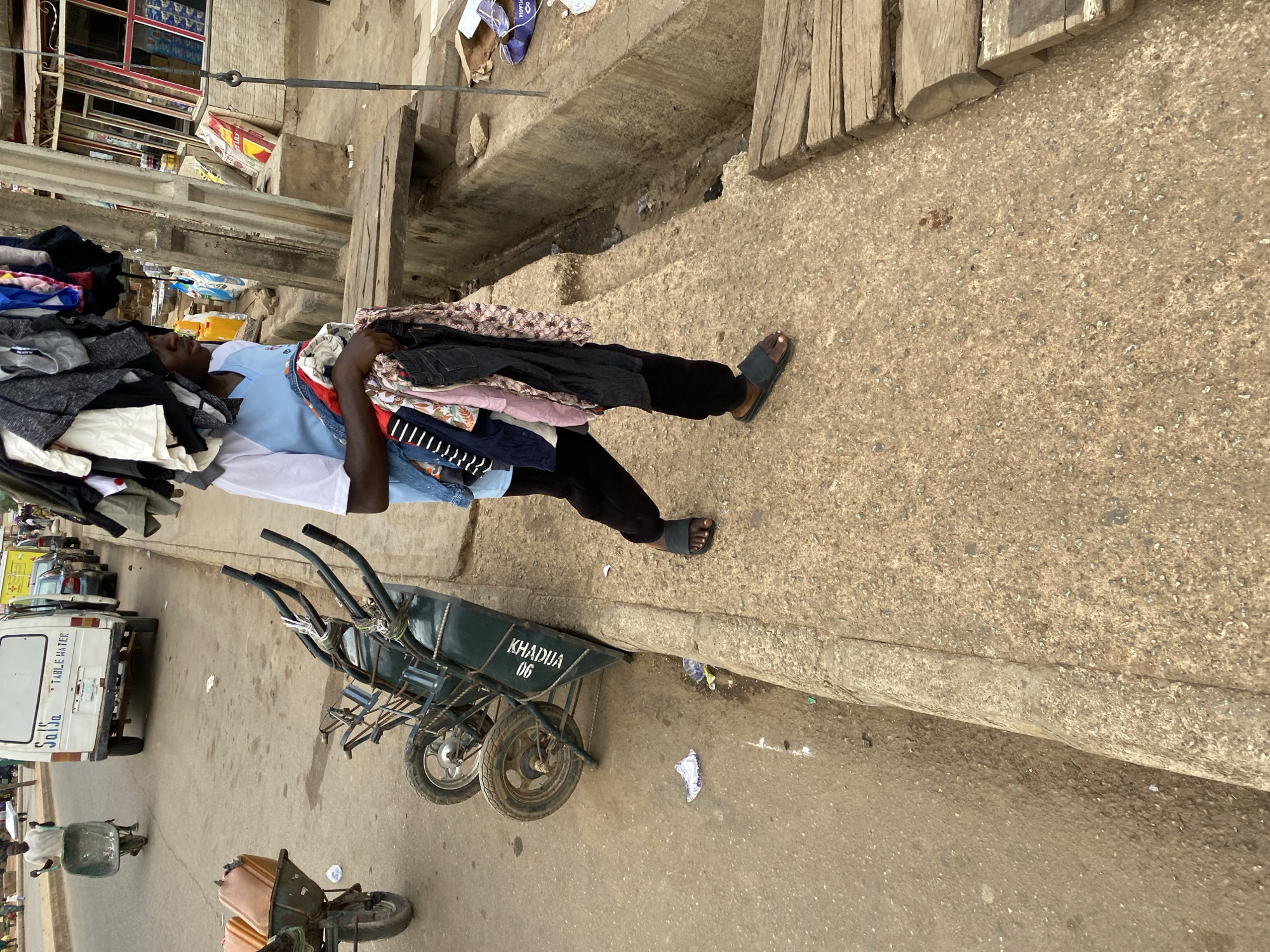 A man carries a large pile of clothes on his head and in his arms while walking on a sidewalk in Bukuru, Nigeria. He is wearing a light blue t-shirt, black pants, and sandals. Next to him are several green wheelbarrows with the name "KHADUA 06" stenciled on them. A white van with the words "TABLE WATER" on the back is parked in the background. The scene appears to be a busy street with various buildings and people visible in the background.