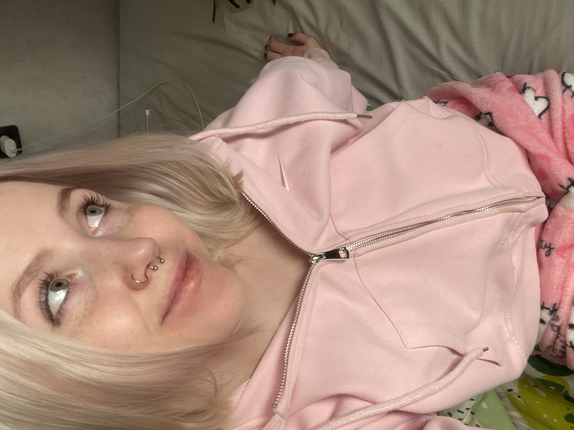 A close-up, high-angle shot depicts a young person lying down, likely on a bed, in an indoor setting. The individual has fair skin, blonde hair, and visible freckles. Their green or light brown eyes are wide and looking upwards, and they wear mascara. Notable facial features include a septum piercing (circular barbell) and a nostril piercing (hoop).
The person is wearing a light pink hooded sweatshirt (hoodie) with a silver zipper, which is partially unzipped. A small, light pink embroidered design, resembling a "swoosh" logo, is visible on the chest of the hoodie. A pink drawstring is also present on the hood. To the right, a soft pink fleece blanket patterned with white hearts and black outlines is draped. A hand with dark nail polish (possibly red or burgundy) rests on the pink hoodie in the upper right.
The background shows plain grey or beige bedding. An electrical outlet with a white plug is visible near the person's head on the left, and a charging cable trails across the bedding. The dim lighting suggests an evening or night setting indoors. The person's expression is relaxed, with a soft, upward gaze. No discernible text is visible.