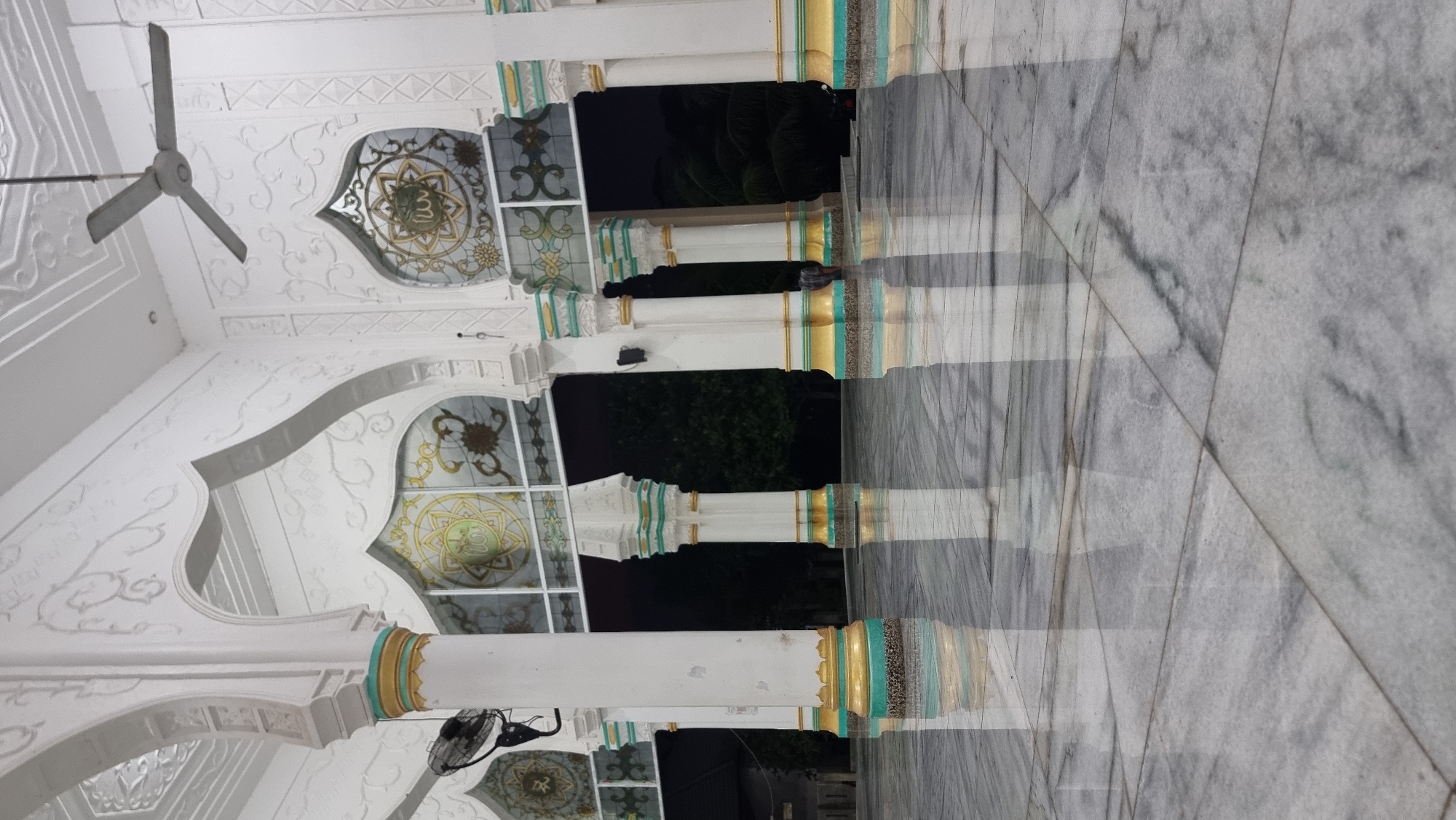 The image shows the interior of a mosque in Lapang Timu, Indonesia. The floor is made of polished white marble with gray veining, reflecting the architectural elements above. The scene is framed by tall, white columns with decorative gold and teal bands near their bases. Above the columns are arched openings with stained glass windows depicting intricate Islamic geometric patterns and calligraphy. A white ceiling with embossed detailing is visible at the top of the frame, and a gray ceiling fan is mounted in the upper right corner. The background shows a dark, out-of-focus view of trees and vegetation, suggesting an outdoor area beyond the mosque's interior. The lighting appears to be artificial, casting reflections on the glossy floor. No people are clearly visible in the foreground, but a person in dark clothing can be seen in the mid-ground, near the center-right of the image, partially obscured by a pillar.