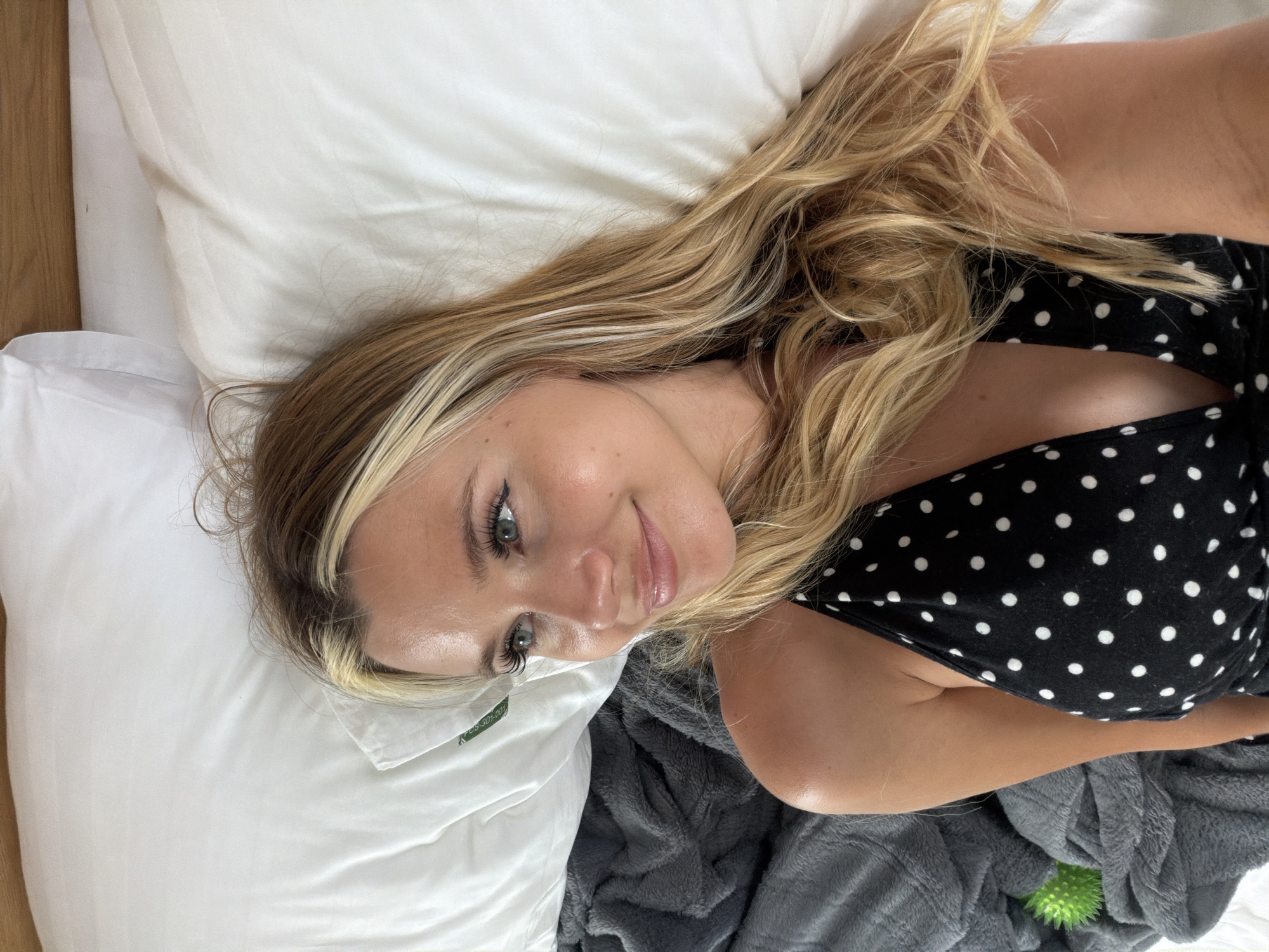 An overhead shot shows a young Caucasian woman lying on a bed with white sheets and a wooden headboard. She has long blonde hair with lighter streaks and is wearing a black halter top with white polka dots. Her face is turned to the side, with her eyes looking upwards. She has on light makeup, including eyeliner and lip gloss. To the left of the bed, a gray, fuzzy blanket is visible, with a small, spiky green ball on it. The lighting suggests it's daytime, possibly in a bedroom in Thep Krasatti, Thailand.