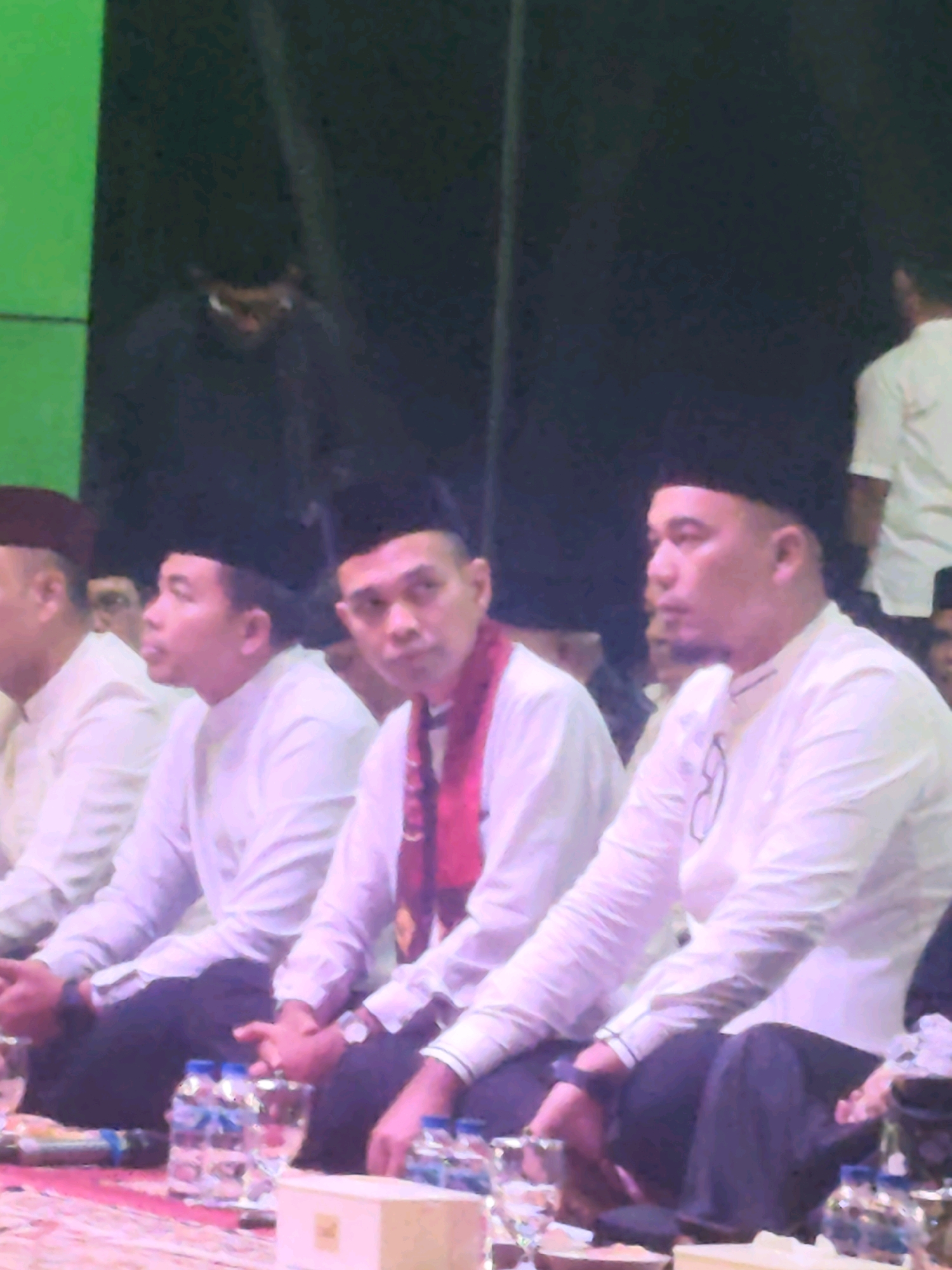 The image depicts a group of men, likely Indonesian, dressed in traditional attire, including peci caps and white shirts. They are seated, with some wearing red scarves. In the foreground, there are bottles of water and glasses on a surface that appears to be a stage or floor. The background is dark, with some subtle hints of a green backdrop on the left side. The overall atmosphere suggests a formal gathering or event. The image does not contain any discernible clues that would place it in the city of Rengat, Indonesia.