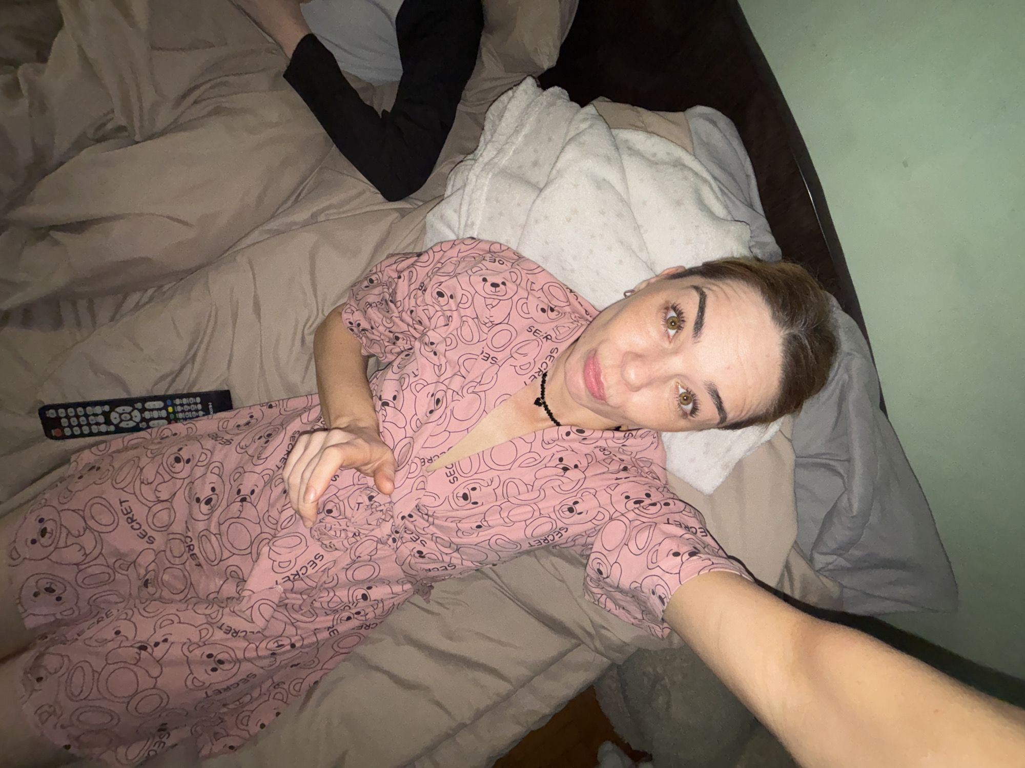 A person with light brown hair and brown eyes lies on a bed with light brown bedding. They are wearing a pink robe with a pattern of teddy bears and the word "SECRET" printed on it. The person is looking up at the camera, with their right arm bent and hand resting on their chest. To the left of the person is a black remote control. To the right of the person, another person's legs are visible, wearing black pants. The scene appears to be indoors, likely in a bedroom. The image was taken in the city of Sinyavino, Russia.