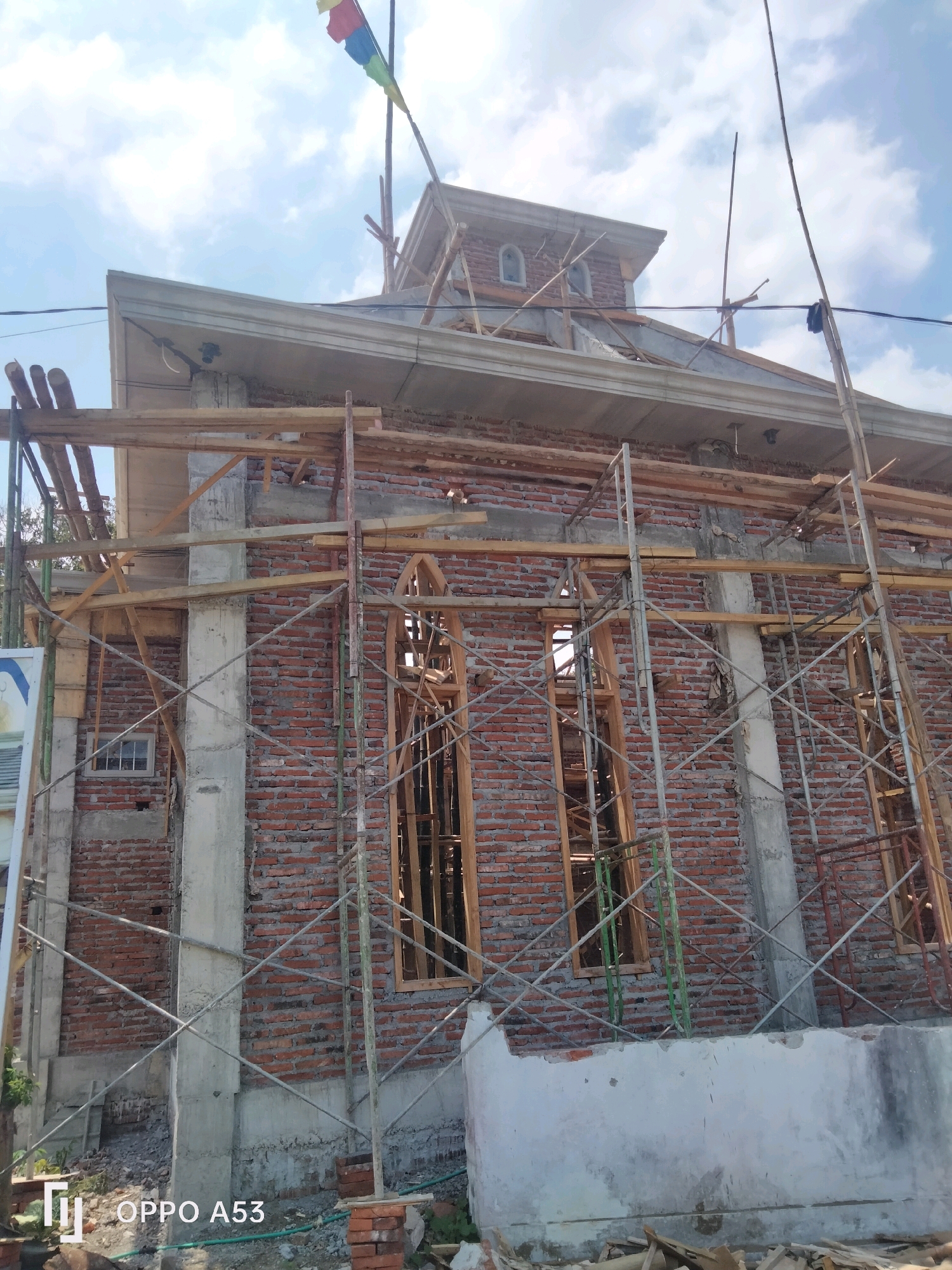 An exterior, low-angle, slightly left-tilted, daylight, medium shot captures a brick building under construction, set against a bright blue sky with scattered white clouds. The building features visible scaffolding made of metal poles and wooden planks. The walls are constructed of red bricks with concrete pillars and window frames. Two arched window openings are visible on the main facade. The roofline is partially visible, with a concrete overhang. A multi-colored flag flies from a pole at the top of the structure. Debris and construction materials are scattered on the ground around the base of the building. The bottom right corner of the image displays the text "OPPO A53" in white font.