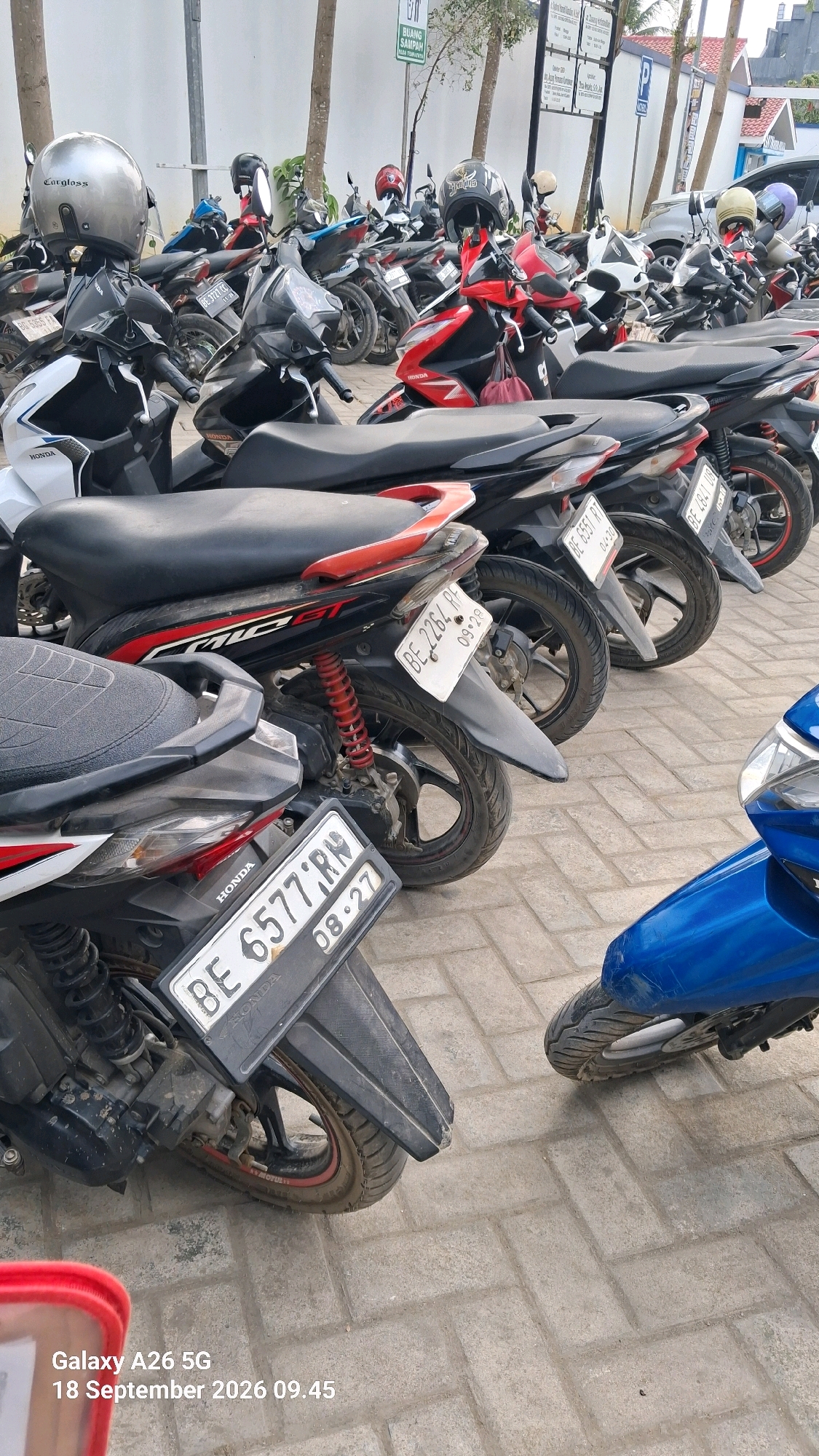 The image depicts a parking area for motorcycles in Kutoarjo, Indonesia. Numerous motorcycles, predominantly of the scooter type, are parked in rows on a brick-paved surface. Some motorcycles are red and black, while others are white and black. Several helmets, including a silver one and a black one with a design, are placed on the handlebars or seats of the parked motorcycles. The environment appears to be outdoors, with trees and buildings visible in the background. Signs are also present, one of which indicates "BUANG SAMPAH" (trash disposal). The timestamp on the image indicates it was taken on September 18, 2026, at 09:45.
