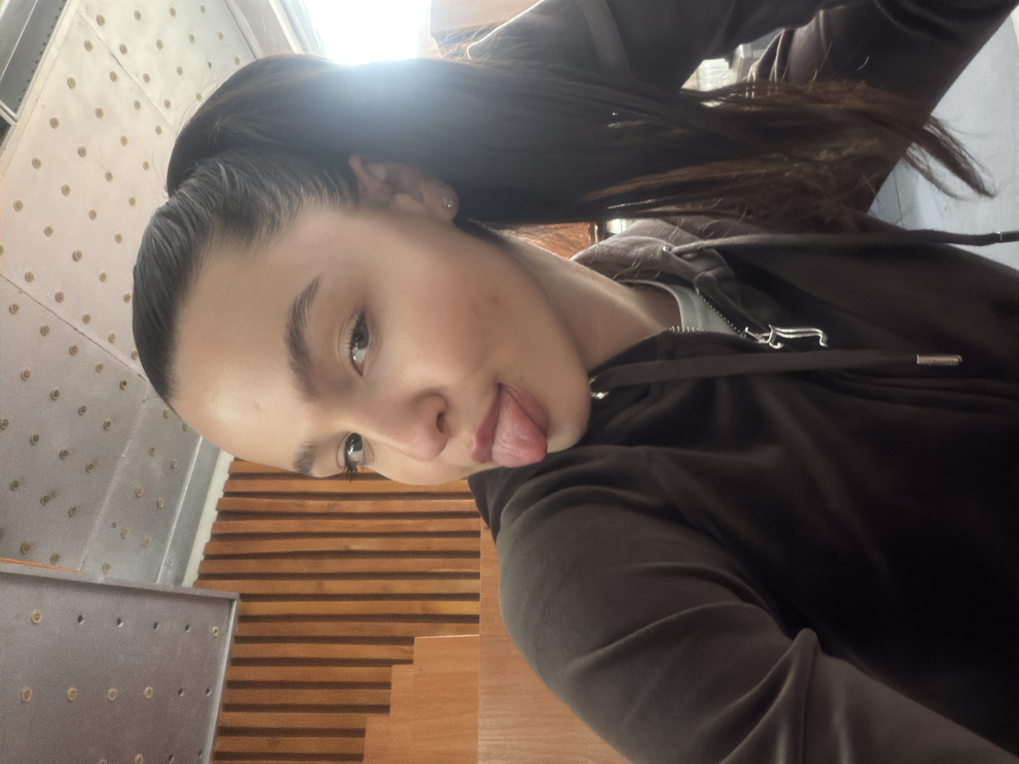 The image, rotated counter-clockwise, presents a close-up selfie of a young individual, likely female, captured indoors in Sofia, Bulgaria. The subject has dark, possibly brown or black, hair pulled back into a high ponytail, with some strands flowing towards the upper right of the frame (when corrected). She is wearing a dark brown or charcoal-colored hooded sweatshirt or jacket made of a soft, possibly velour or fleece, material, with a zipper down the front and a glimpse of a light-colored undergarment at the neckline. A small stud earring is visible in her left ear.
Her face is tilted slightly towards the viewer, with her left eye, which appears light-colored, clearly visible. She is playfully sticking out her tongue, conveying a cheeky and casual demeanor. Faint blemishes are present on her right cheek.
The background reveals distinct architectural elements. To the left of the subject, a light-colored, possibly metallic, wall or panel is visible, patterned with a regular arrangement of small, dark circular indentations, resembling rivets or decorative studs. To her right and below, there is a wooden surface featuring parallel horizontal stripes, suggesting stairs, slatted wall paneling, or flooring. A partial view of a metal railing or barrier is also discernible in the lower-left foreground. Bright light emanating from the upper background indicates a daytime setting, likely near a window or skylight. No discernible text is visible in the image. The scene conveys a lighthearted, informal moment within a contemporary interior.