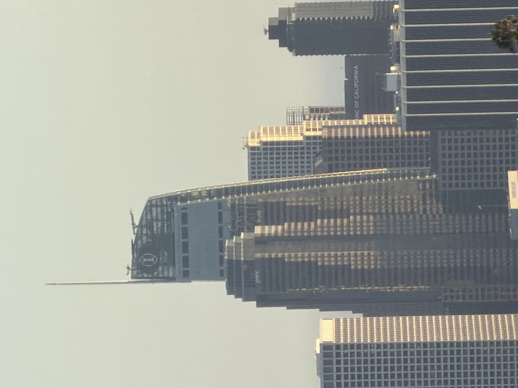 This is a medium shot of a cityscape at what appears to be sunrise or sunset, with the sky a pale yellow and white. The scene is dominated by several skyscrapers. In the foreground and center is a tall, dark building with many recessed windows and a unique, slanted top that features a spire and a large circular logo with an "I" inside. The building also has "pwc" written on its side in white lettering. To the left of this building is a lighter colored skyscraper with a grid-like pattern of windows. To the right of the central building, there are other skyscrapers, including one dark, rectangular building with a flat roof, and a building with a sign that reads "ANC OF CALIFORNIA". The overall impression is of a dense urban environment.