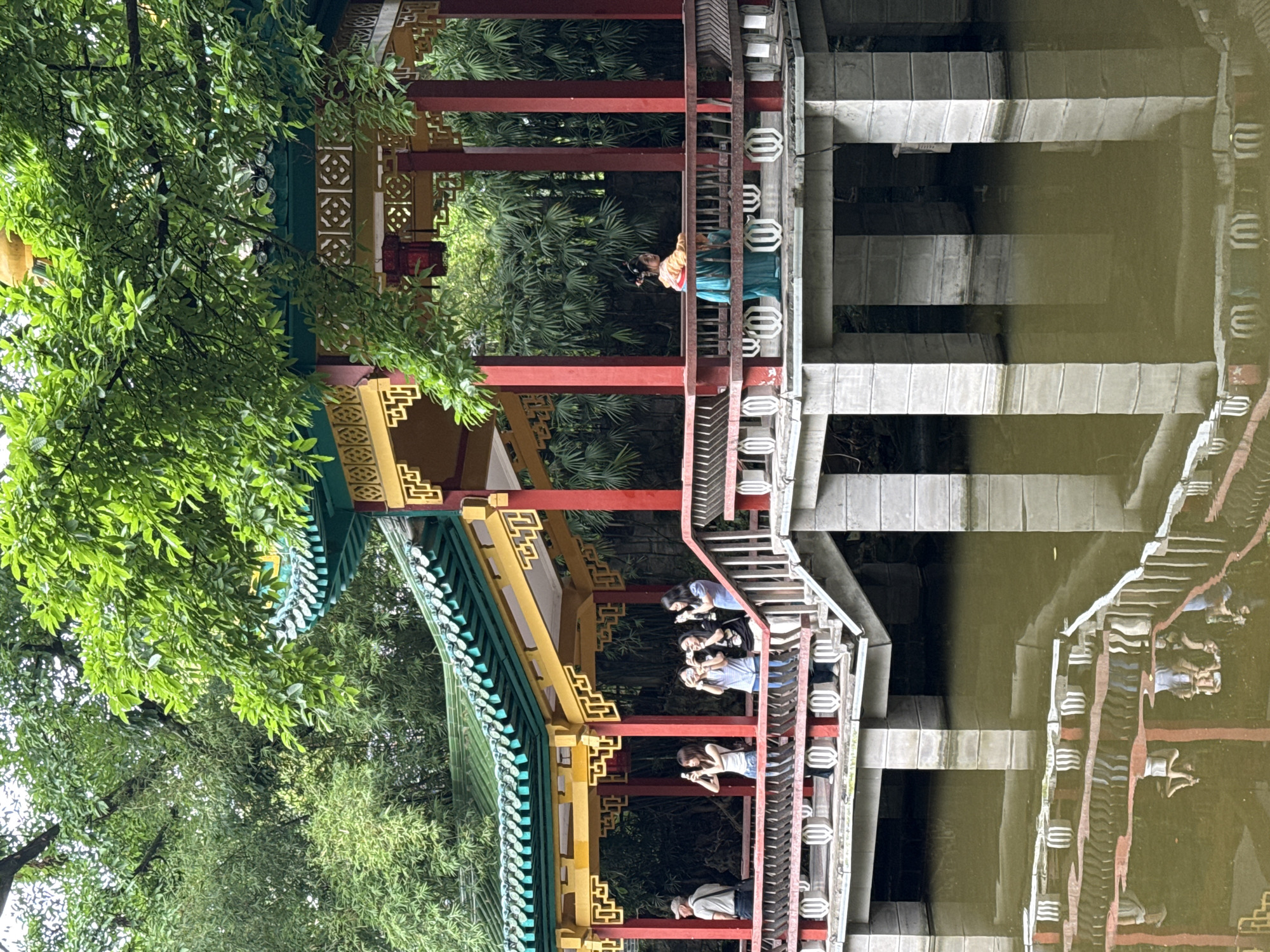 The image shows a pavilion structure on the edge of a body of water, likely a lake or pond, with its reflection visible in the water. The pavilion is decorated in traditional Chinese style with red columns and ornate yellow and green eaves. Several people are visible on the pavilion, some taking photographs with their phones. Another person wearing traditional Chinese clothing stands separately on the structure. The scene is likely in the Yuzhong District of Chongqing, China, based on the traditional architectural style. A tree with green leaves is located on the left side of the image.