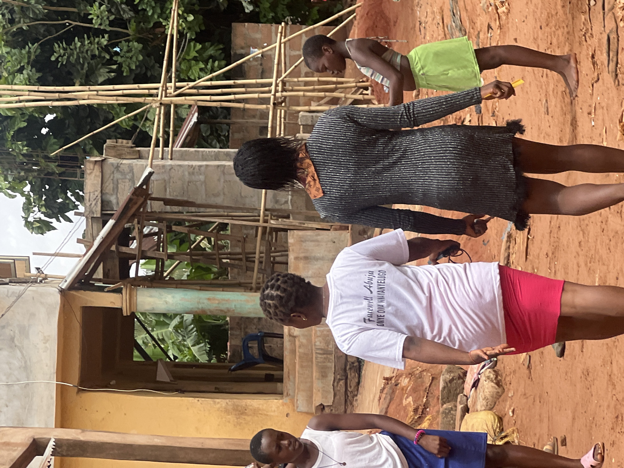 The scene appears to be in Onitsha, Nigeria, showing a group of people in an outdoor setting. The environment consists of unfinished buildings and a bamboo scaffolding structure.
A woman with braided hair, wearing a grey, striped dress, is holding a yellow object in her right hand. Near her is another young woman wearing a light green dress with a white top. Standing near to the first woman is another woman wearing a white t-shirt with the words "Farewell Abuja Onye Oma Nwanyelugo" and a red skirt. In the foreground, a young woman lies on the ground, wearing a white t-shirt and a blue skirt. The ground is red and dusty.