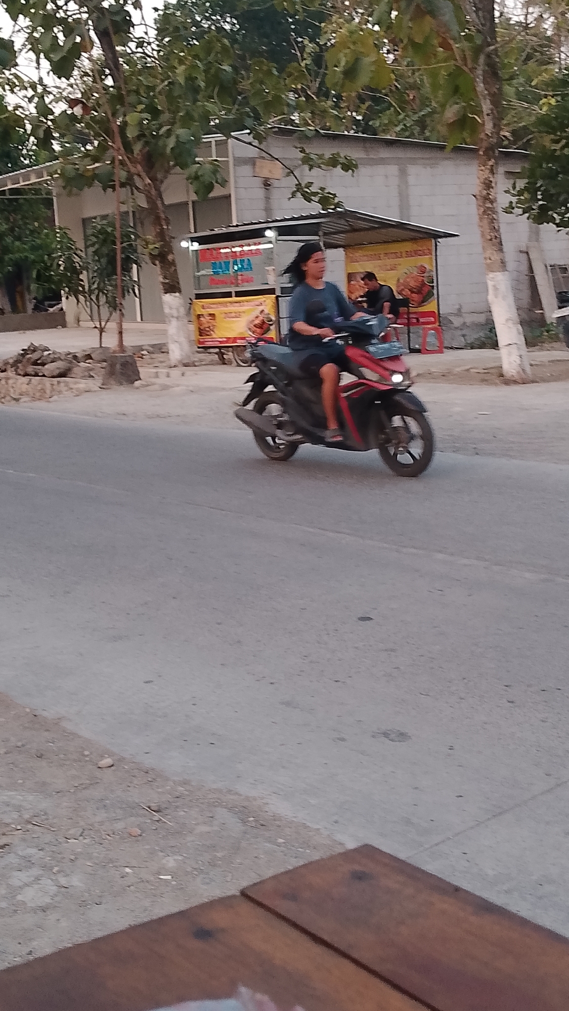 A woman with dark hair rides a red and black scooter on a paved road in Karanglangu, Indonesia. She wears a dark blue t-shirt and dark shorts. The scooter has a silver exhaust pipe and a license plate that reads "AB 3456". In the background, there is a building with a white roof and gray walls, a small food stall with yellow and red signage, and several trees with green leaves. The road is gray asphalt, and there is a dirt and gravel area to the left of the road. A wooden table is visible in the foreground.