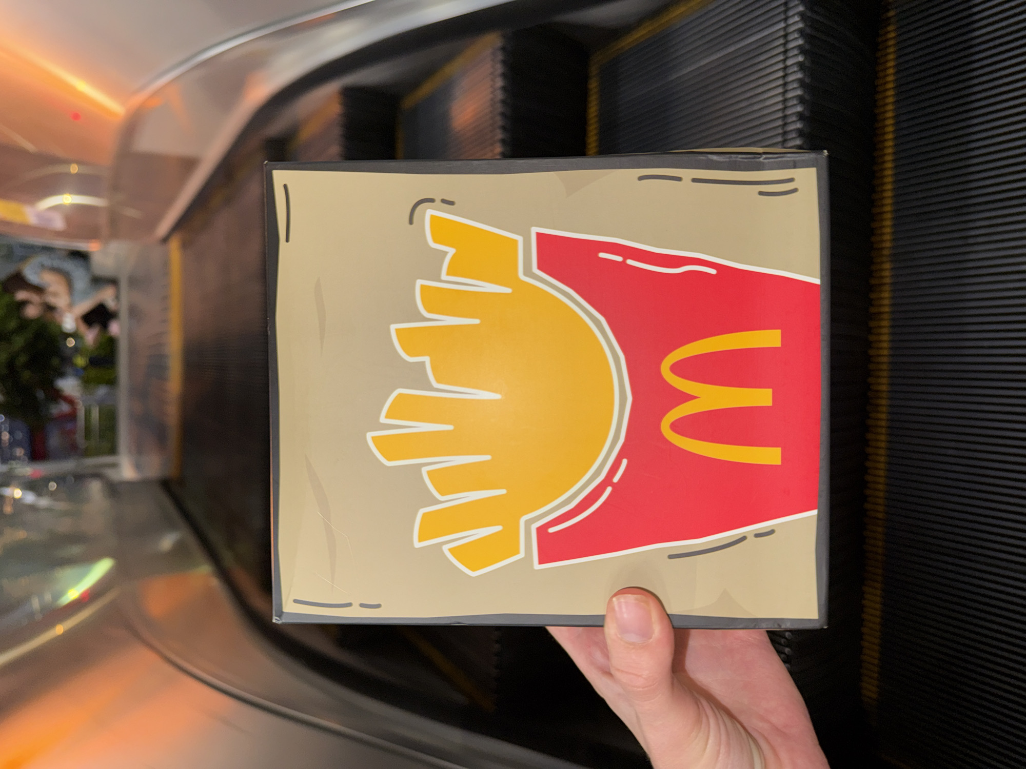 A person's left hand holds a square cardboard box with a cartoon drawing of McDonald's french fries and a logo on it. The box is placed in front of an escalator. The escalator steps are black with yellow lines. In the background, two blurred figures are seated on a bench. The scene appears to be in a public place, possibly a mall or transportation hub, in Nanshan District, China. The lighting suggests an indoor environment with artificial illumination.