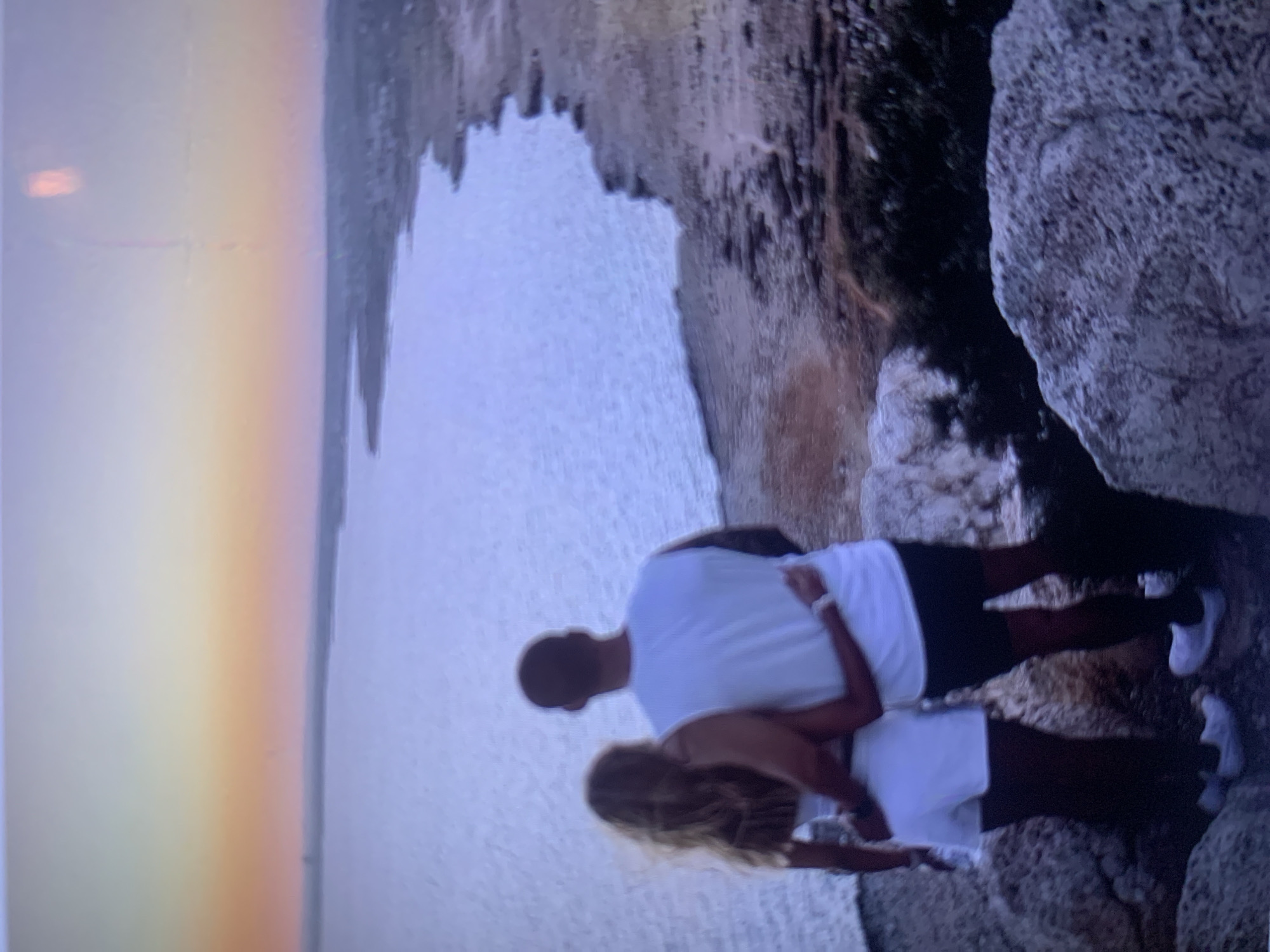 The image shows a couple embracing on a rocky cliff overlooking a body of water at sunset. The man, wearing a white t-shirt and dark shorts, has his arm around the woman, who is wearing a white top and white shorts. The sun is low in the sky, casting a warm orange and pink glow on the horizon. The water is calm and reflects the colors of the sunset. The coastline is visible in the background, with some vegetation and buildings. The couple appears to be enjoying the view. This location is likely the Black Sea coast near Burgas, Bulgaria, possibly a cliff area like those found near the city.