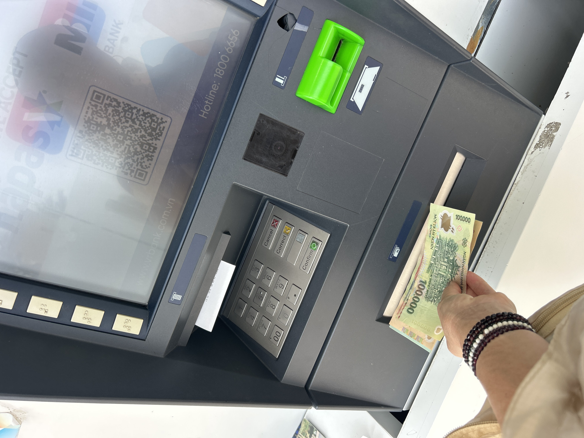 A high-angle shot depicts an ATM in what appears to be Bắc Nha Trang, Vietnam, based on the Vietnamese currency and language on the ATM. The ATM is gray, with a keypad displaying options in English. A hand wearing multiple beaded bracelets holds Vietnamese 100,000 Dong banknotes emerging from the cash dispenser. The screen is partially visible, showing Vietnamese text and a QR code. The ATM also features braille markings, a card slot with a green guard, and a paper dispenser. The ATM itself has some wear and tear, with visible scuffs and marks. The overall scene suggests a person withdrawing money from an ATM.