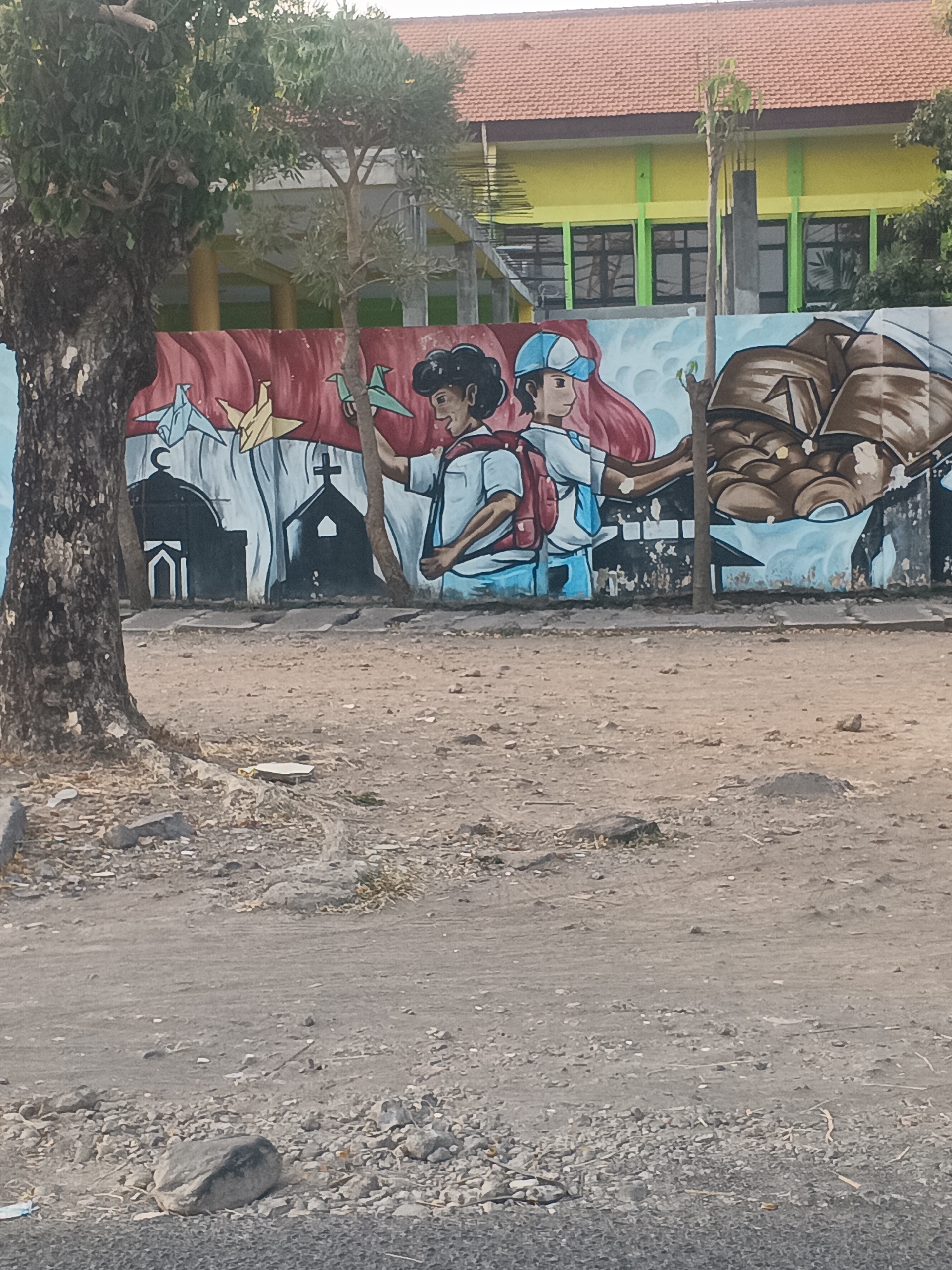 The image shows a mural on a wall in front of a building with a yellow facade and a red tiled roof, likely a school in Surabaya, Indonesia. The mural depicts two students, one wearing a blue cap and the other with dark hair, holding origami birds. To the left of the students, there are silhouettes of religious buildings, including a mosque with a crescent moon and a church with a cross. To the right, the mural shows an abstract representation of earth or rocks. A large tree stands to the left of the mural, and the ground in front of the wall is covered with dirt and gravel, indicating an outdoor urban environment.