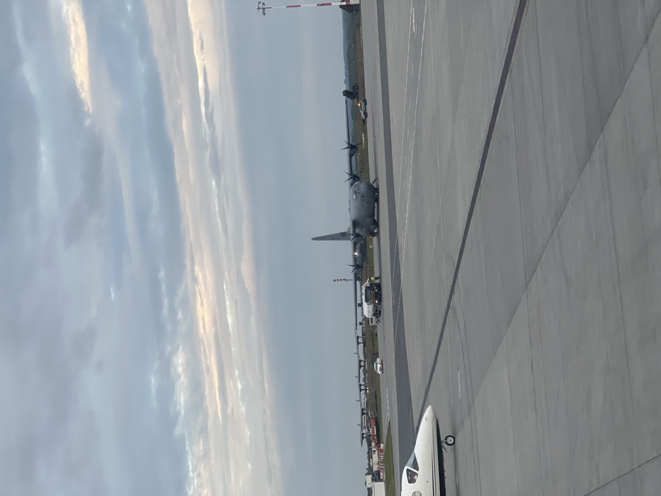 The image shows a large gray military aircraft, likely a Lockheed C-130 Hercules, parked on the tarmac of the Jasionka Airport in Jasionka, Poland. The coordinates provided correspond to the airport's location. A white private jet is parked closer to the camera. The sky is cloudy. A fuel tanker truck is parked behind the military aircraft. Several figures appear to be interacting around the large aircraft, possibly maintenance personnel. The aircraft is facing away from the camera, and its tail is in the center of the frame. The image is taken from a low angle, showing the runway and the aircraft's front landing gear. There are lines painted on the runway.