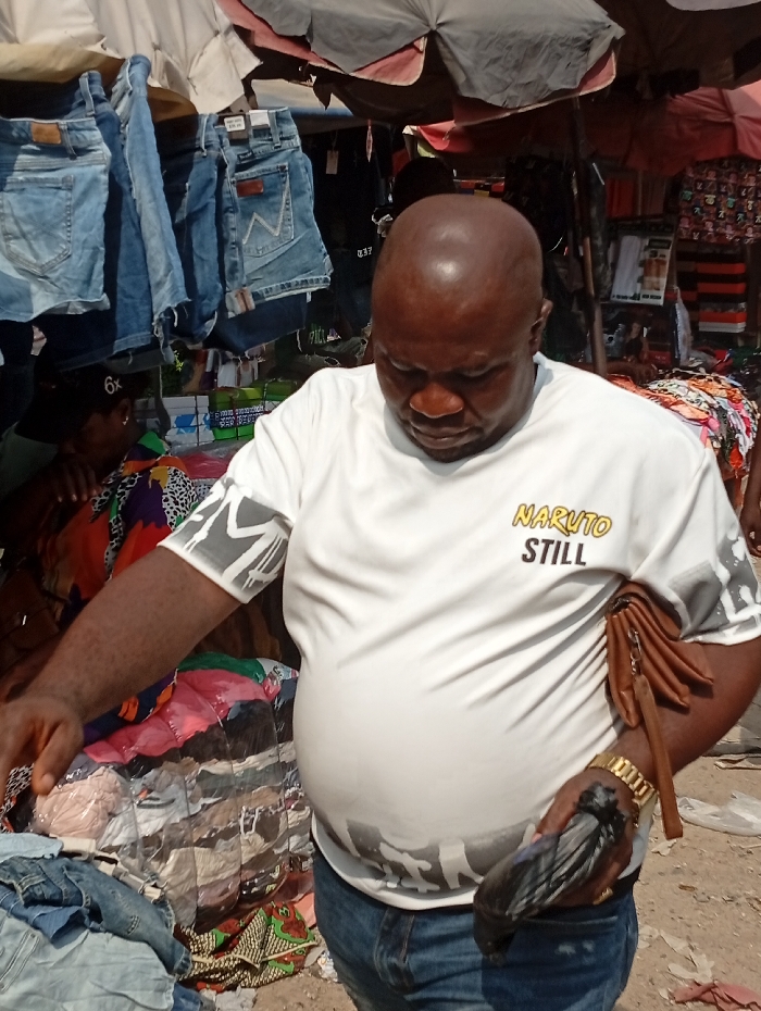 A bald man in a "Naruto Still" T-shirt shops at an outdoor market. He is examining a pile of clothes, with denim items nearby. A brown wallet hangs from his side. Other merchandise, including jeans and packaged items, are visible in the background. An umbrella provides shade. The man is also wearing a gold watch.
