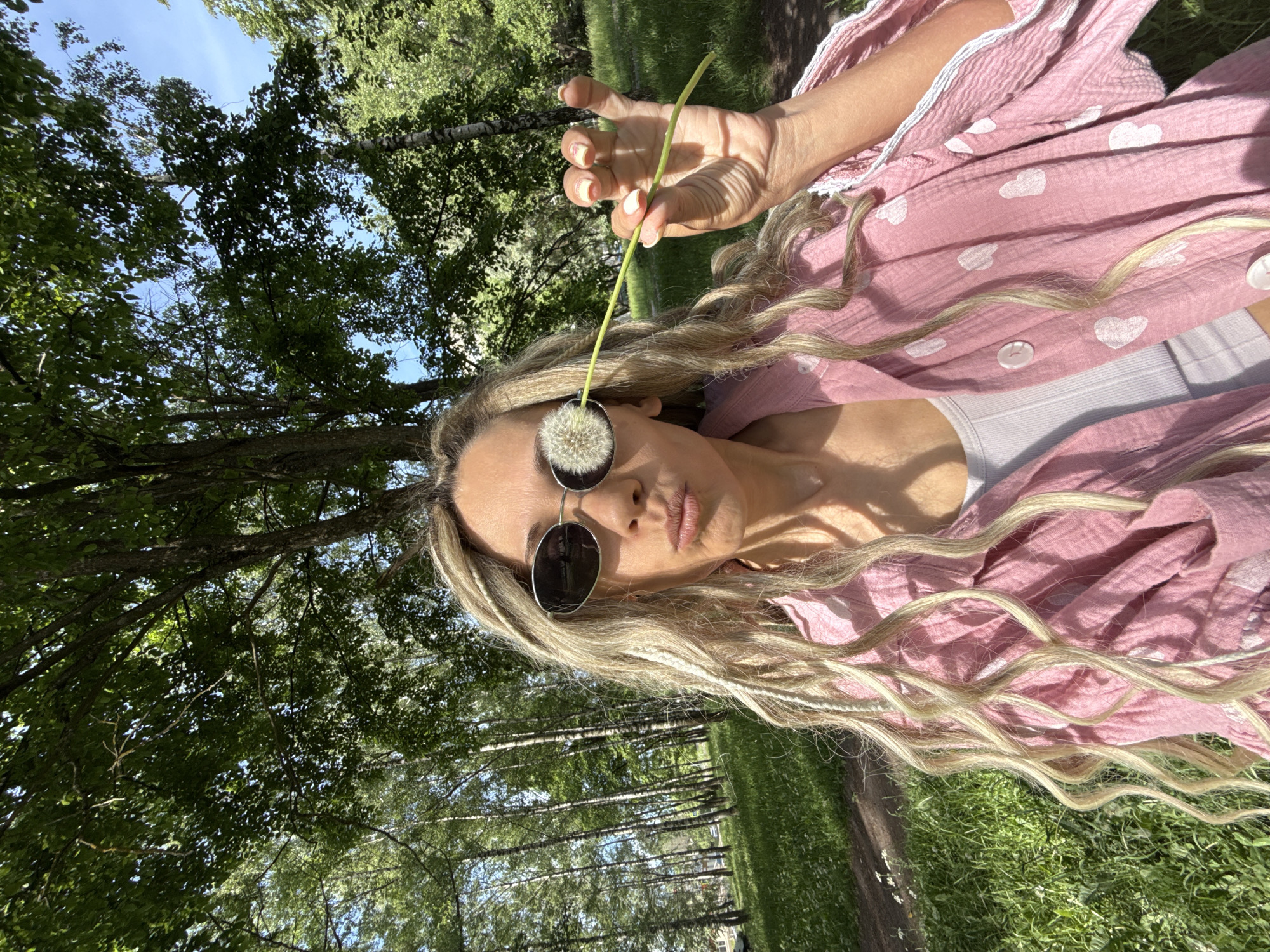 A forensic, matter-of-fact description of the scene reveals a lone adult female outdoors in a natural setting. The individual is positioned centrally, viewed from a slightly low angle, captured in a close-up. She has long, light-colored hair styled in dreadlocks or braids, with some strands flowing loosely. She wears dark, oval-shaped sunglasses. Her mouth is pursed in a pouting expression. A mature dandelion seed head on a green stem is held by her left hand (viewer's right) in front of her right sunglass lens, obscuring it. She is dressed in a loose-fitting, light-pink, textured shirt or jacket adorned with white heart patterns, open to reveal a light-colored, ribbed crop top underneath. The background consists of dense green foliage, primarily trees, with several distinctive white-barked birch trees visible on the left. Patches of clear blue sky are discernible through the leaves. The ground is covered with lush green grass, and a dirt path curves through the scene on the left side. The lighting is bright and natural, indicating daylight. The specific city and country are unknown, but the location is consistent with a park or natural recreational area.