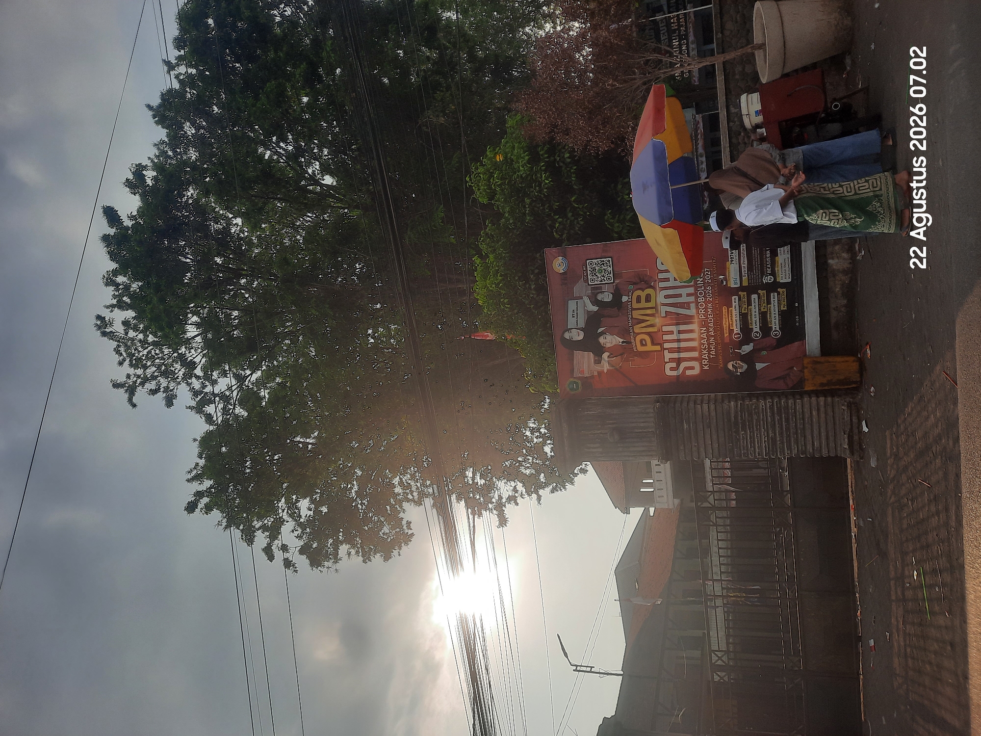The image depicts an outdoor scene under a cloudy sky with the sun partially visible, creating lens flare. In the foreground, a black metal gate is visible, leading into what appears to be a property with a building behind it. To the right of the gate, a large banner is displayed on a post. The banner advertises "PMB STIH ZAHRA" with text that appears to be in Indonesian, indicating it's for student admissions. Below the banner, a small street vendor's stall with a colorful umbrella is set up. Three individuals are standing near the stall, one wearing a white shirt and green sarong, another in a light-colored long-sleeved shirt and blue pants, and a third person wearing a hijab and blue pants. They appear to be interacting or waiting at the stall. Scattered debris, including what looks like discarded paper and plastic, is present on the ground in front of the gate and stall. A street lamp is visible in the background to the left. The overall impression is a street scene in front of a building with an educational institution's promotional material and a small vendor. The timestamp "22 Agustus 2026 07.02" is superimposed on the bottom right corner of the image, suggesting a date and time. The location is identified as Pajarakan Kulon, Indonesia, which is consistent with the Indonesian language used on the banner.