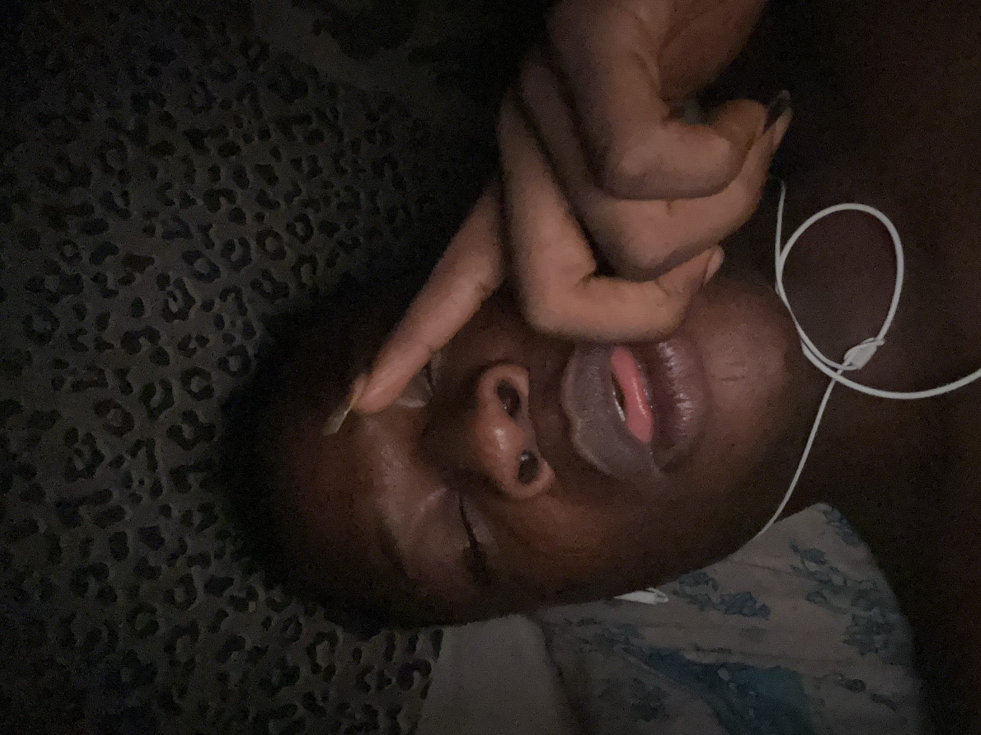 A close-up, low-angle, and slightly tilted image shows a dark-skinned person resting on what appears to be a patterned surface, possibly a bed or couch. The person has their eyes closed, and their hand is positioned over part of their face, with a finger extended towards the forehead. A clear lip mask or balm is visible on their lips. White earbud headphones are near the neck. The setting appears to be indoors, possibly a home, with a dark background. The location is Unknown, Unknown.