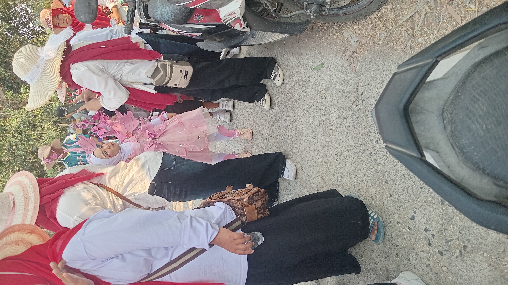 In the city of Kedungwaru, Indonesia, a group of people are gathered outdoors, possibly for an event. A young girl in a pink fairy costume with butterfly wings is the central focus, looking towards a person holding a phone. Several adults, dressed in white shirts and red headscarves or scarves, surround her. One adult is actively taking a photo of the girl with their phone. Another adult, on the left, holds a clear plastic bottle. A motorcycle is parked on the right side of the frame, partially obscuring the view. The ground is a paved surface with scattered leaves and debris. The background features foliage and other people, suggesting a public gathering.