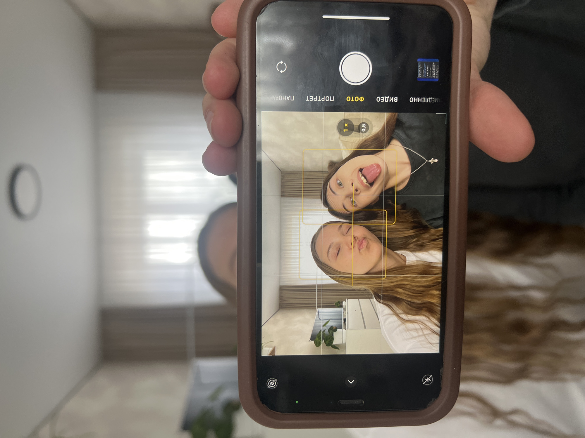 A person is holding an iPhone with a brown case, displaying the camera app interface with a live preview of two young women posing. The woman on the top is sticking her tongue out, while the one on the bottom is puckering her lips as if kissing. The camera is set to "PHOTO" mode with 1x zoom, and grid lines are visible on the screen. The background in the phone's preview shows a room with light-colored walls, a plant, and what appears to be a modern interior design. The wider background outside the phone is blurred, suggesting it is nearby, but not identifiable as being in Votkinsk, Russia. The person holding the phone is partially visible and seems to have long hair.
