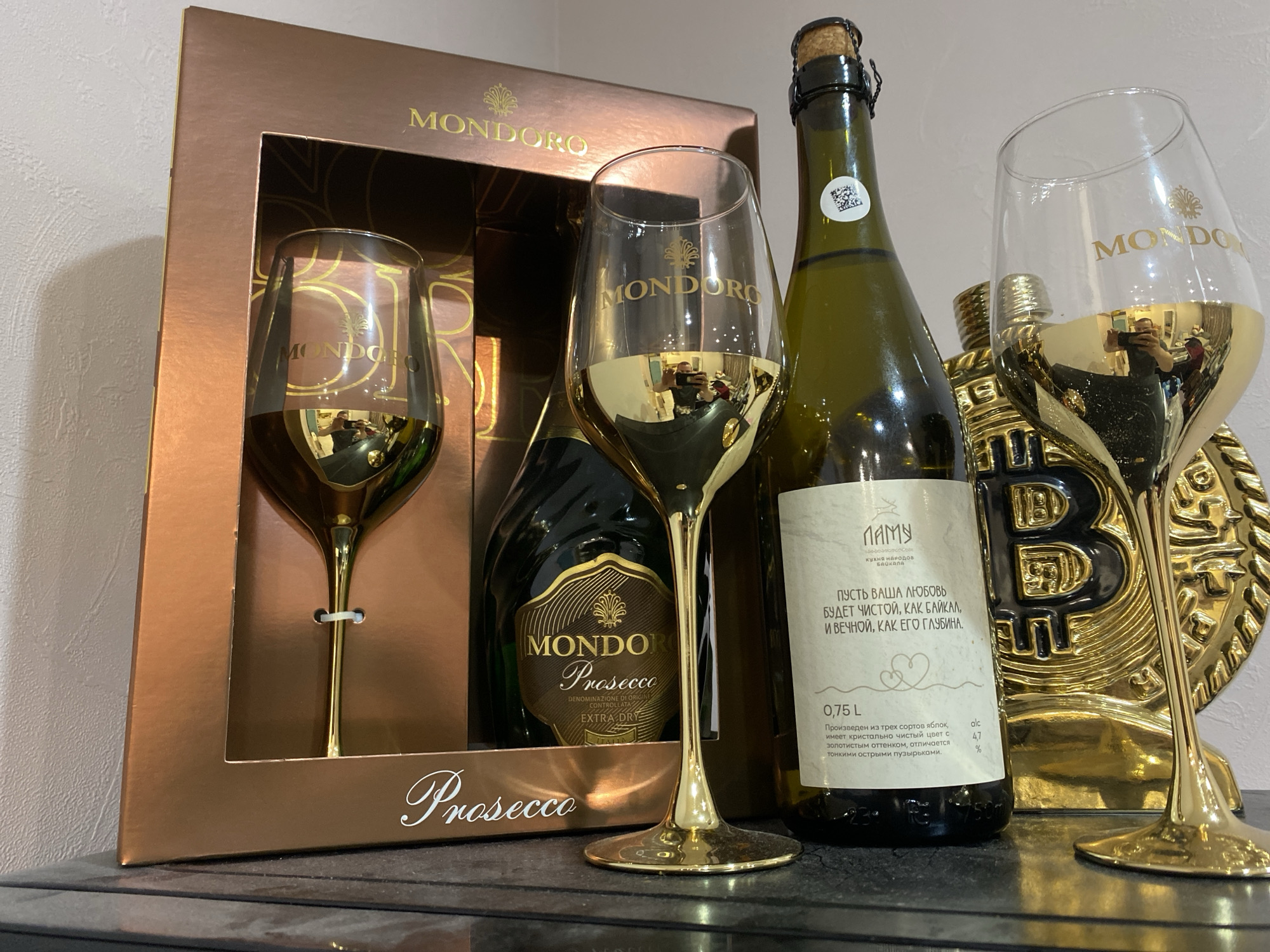 The image shows a still life arrangement of alcoholic beverages and decorative objects. On the left, there's a gift box of "MONDORO Prosecco" which includes two gold-stemmed champagne glasses, one of which is positioned outside the box in the center. Next to the box, there's a bottle labeled "ПАМУ" with text in Russian. To the right, another champagne glass stands beside a golden Bitcoin statue. The background is blurred, suggesting a domestic interior.